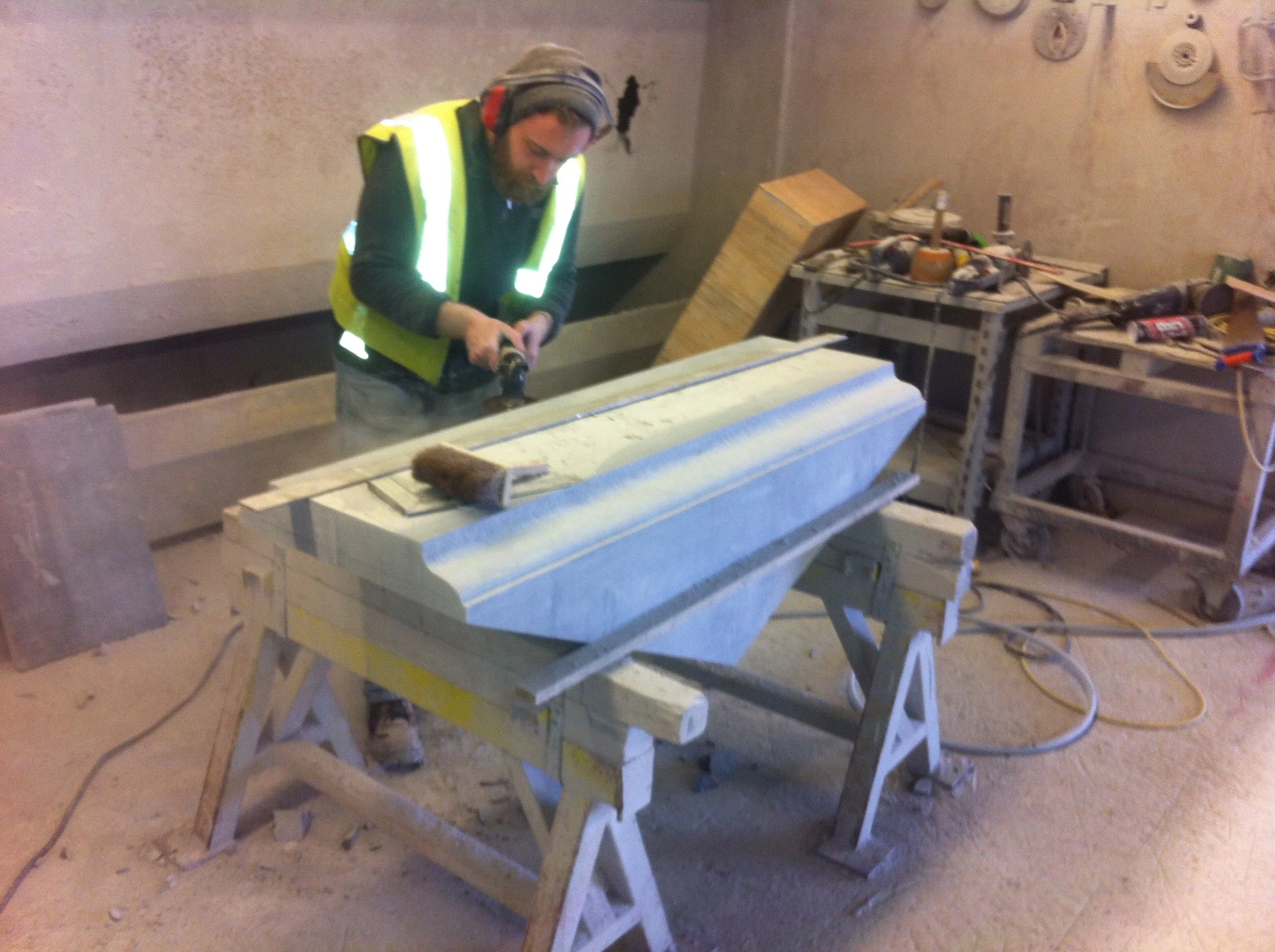 A person working on a woodworking project, wearing a yellow safety vest, in a workshop with tools and materials around.