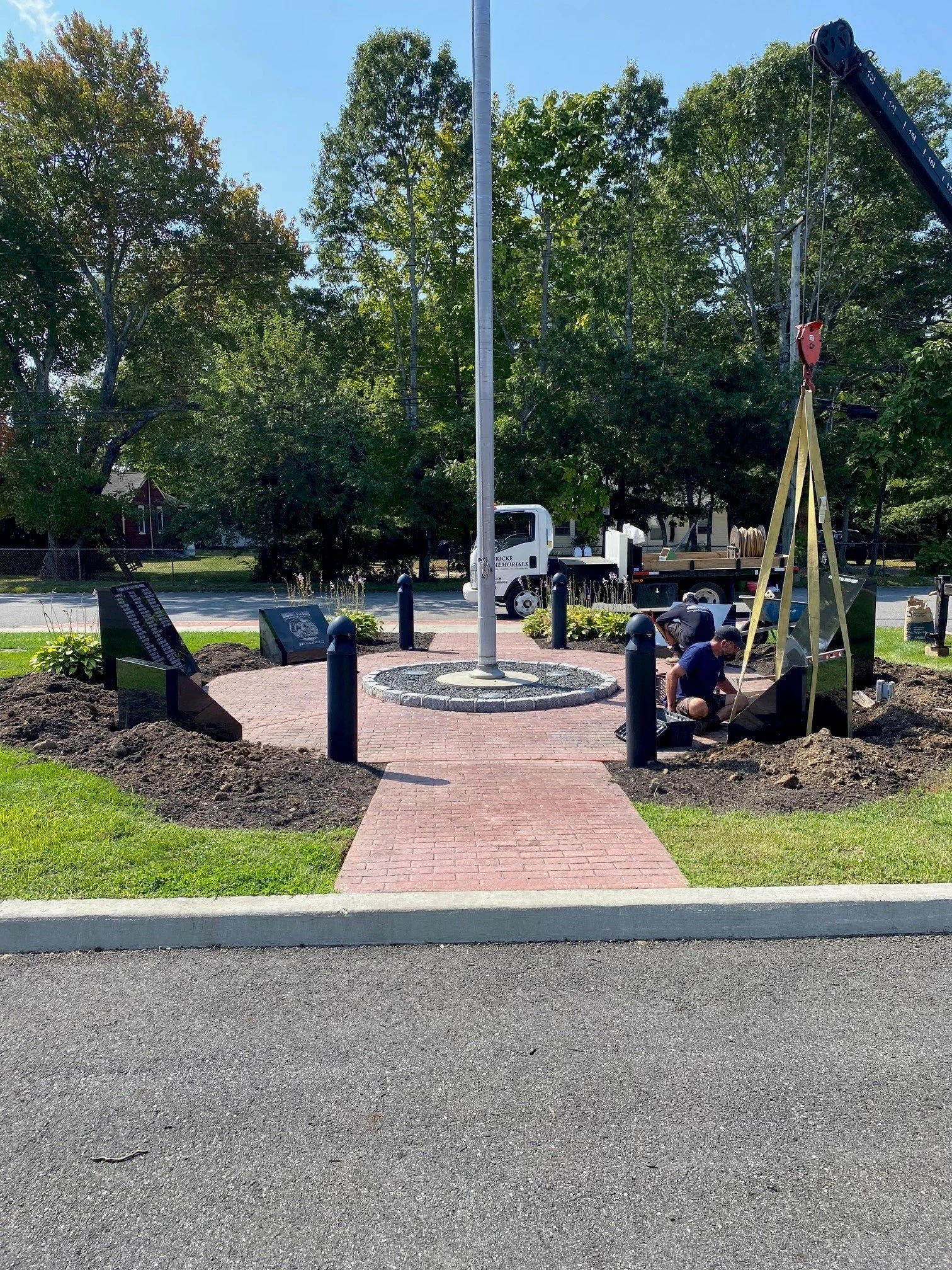 A flagpole with no flag in a small round brick-paved area, flanked by newly planted flowers and shrubs, with workers installing a memorial at the base. In the background, a tree-lined street with a white utility truck, street poles, and green trees.