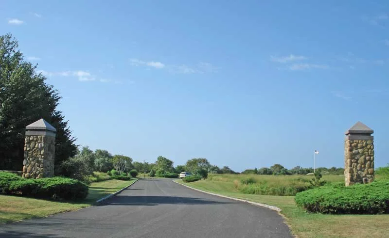 Entrance road to a golf course with stone pillars and well-manicured greenery under a blue sky.