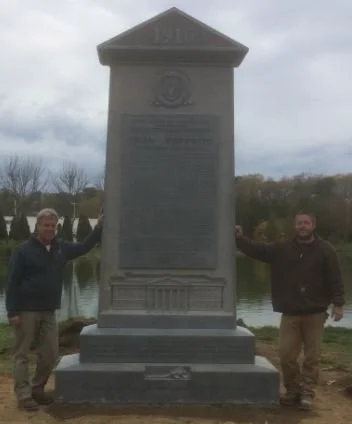Two men standing on either side of a large historical monument by a water body, with trees and cloudy sky in the background.