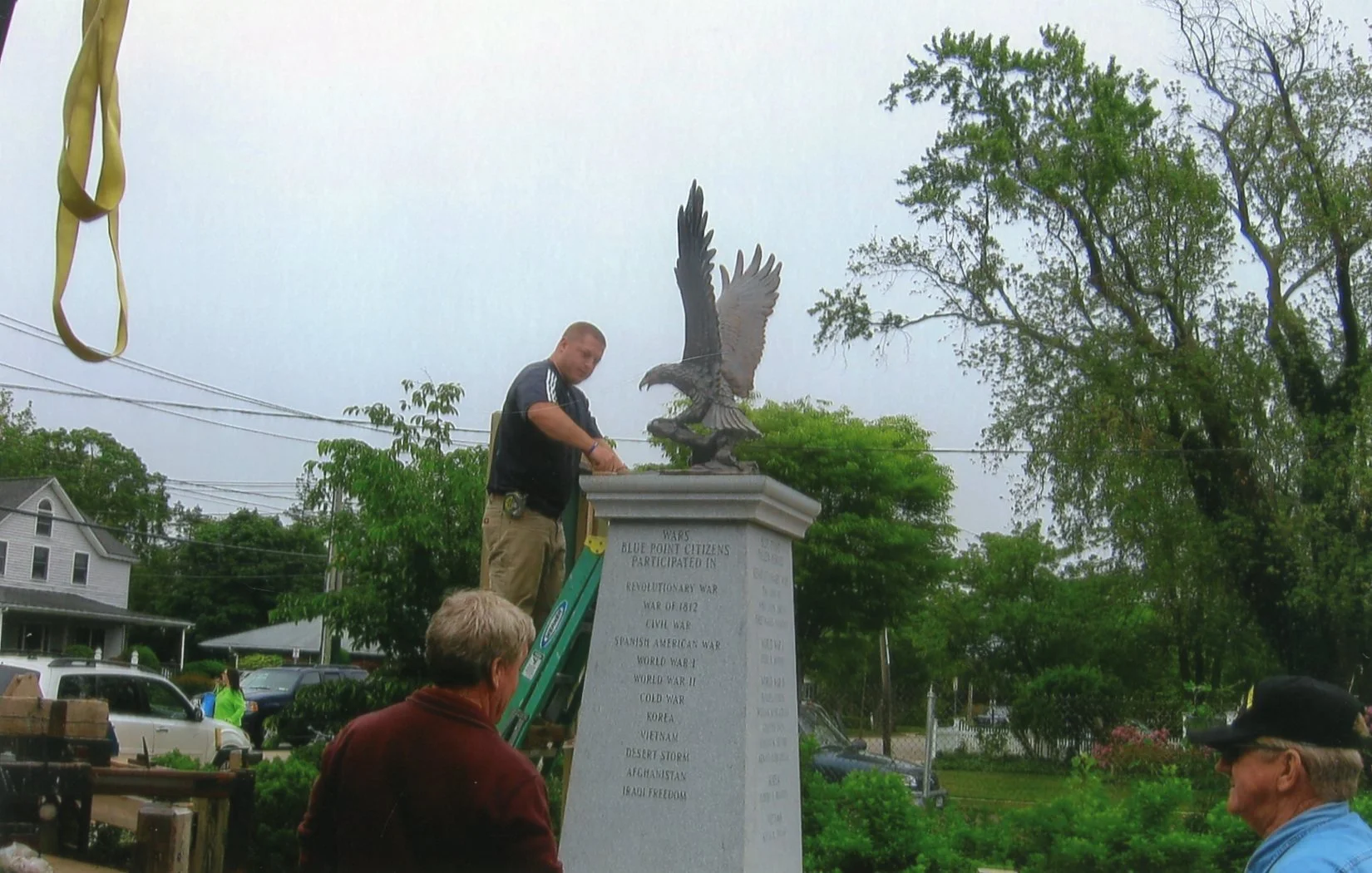 A man on a ladder placing a plaque or wreath on a war memorial with an eagle sculpture on top, in a park-like setting with trees and people nearby.