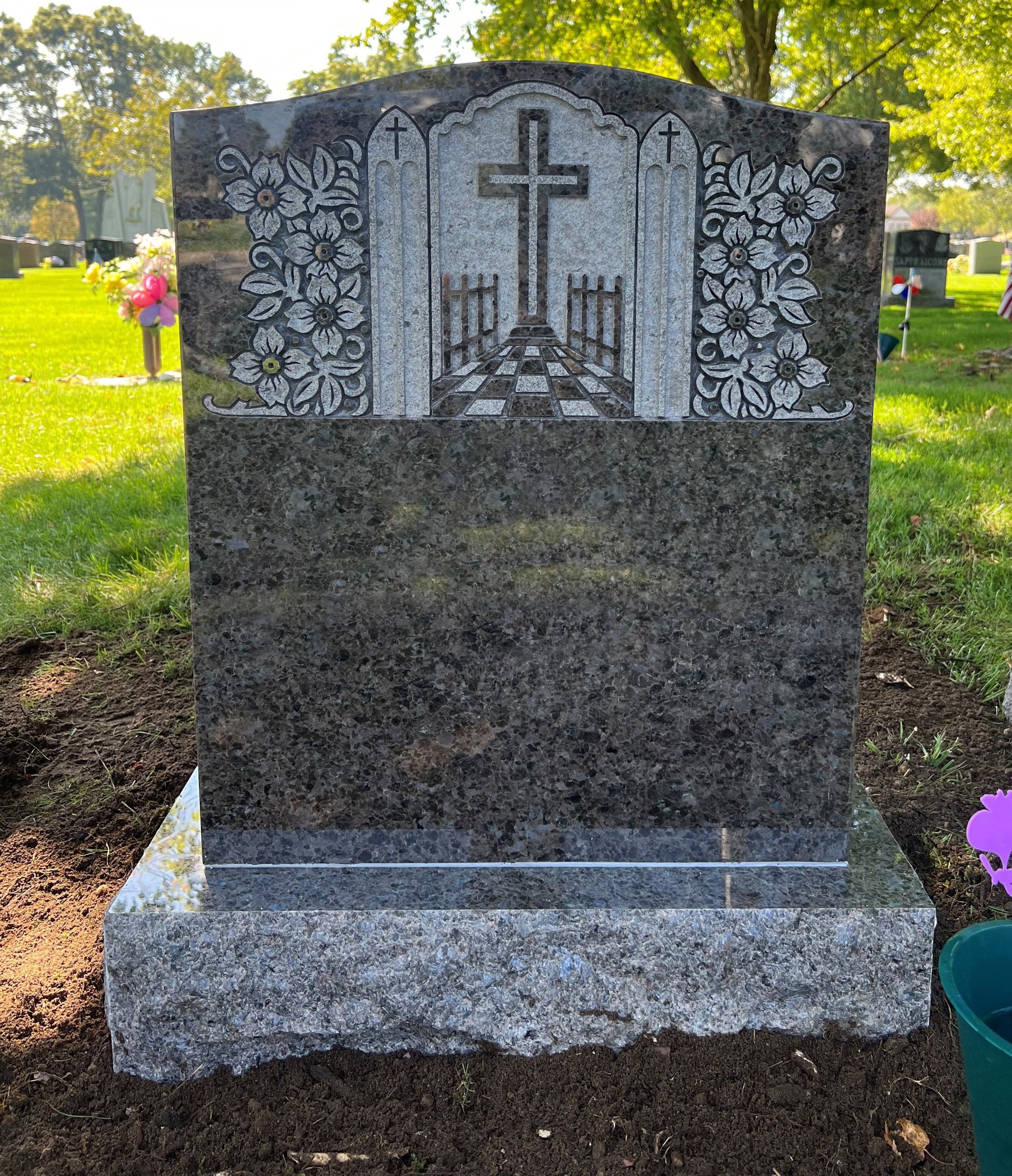 Close-up of a granite headstone with a carved cross, flowers, and gate motif, in a cemetery with trees and other graves in background.