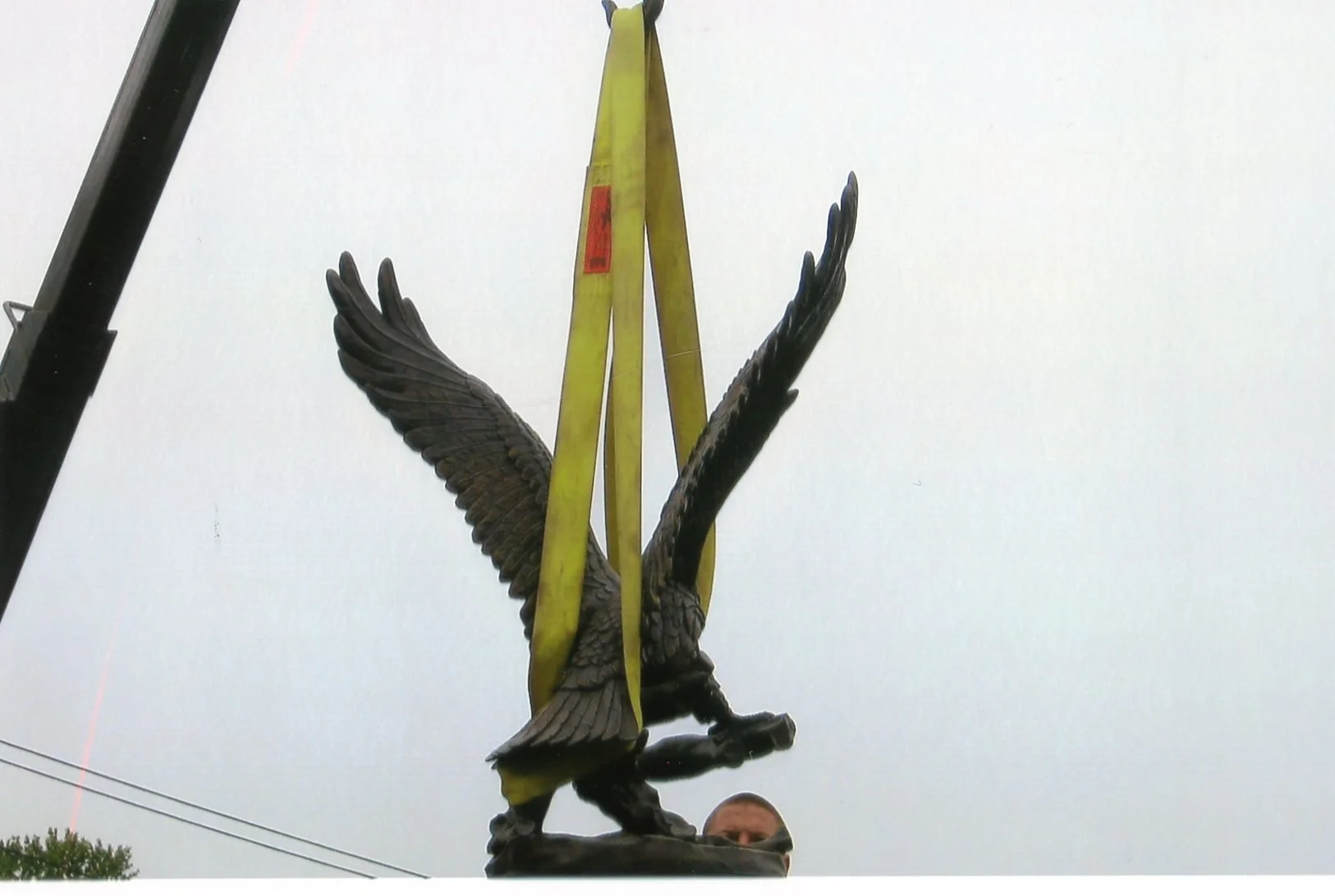 Statue of an eagle with wings spread, mounted on a stand, with a yellow construction lift nearby and a person’s head visible at the bottom of the image.