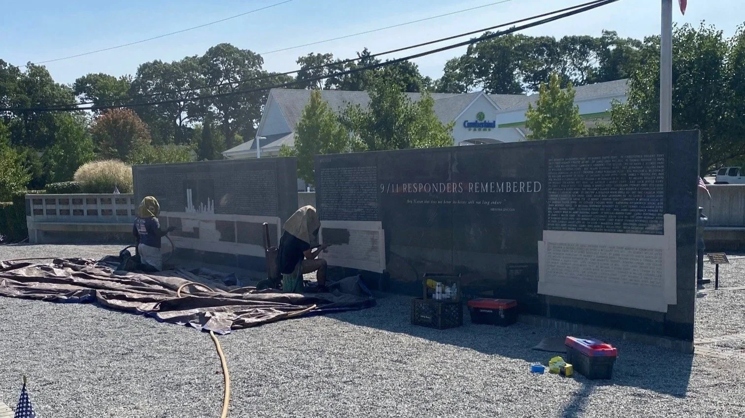 Two people kneeling in front of a memorial wall that displays text and images, with gardening tools and a tarp on the gravel ground, and a road, trees, and buildings in the background.