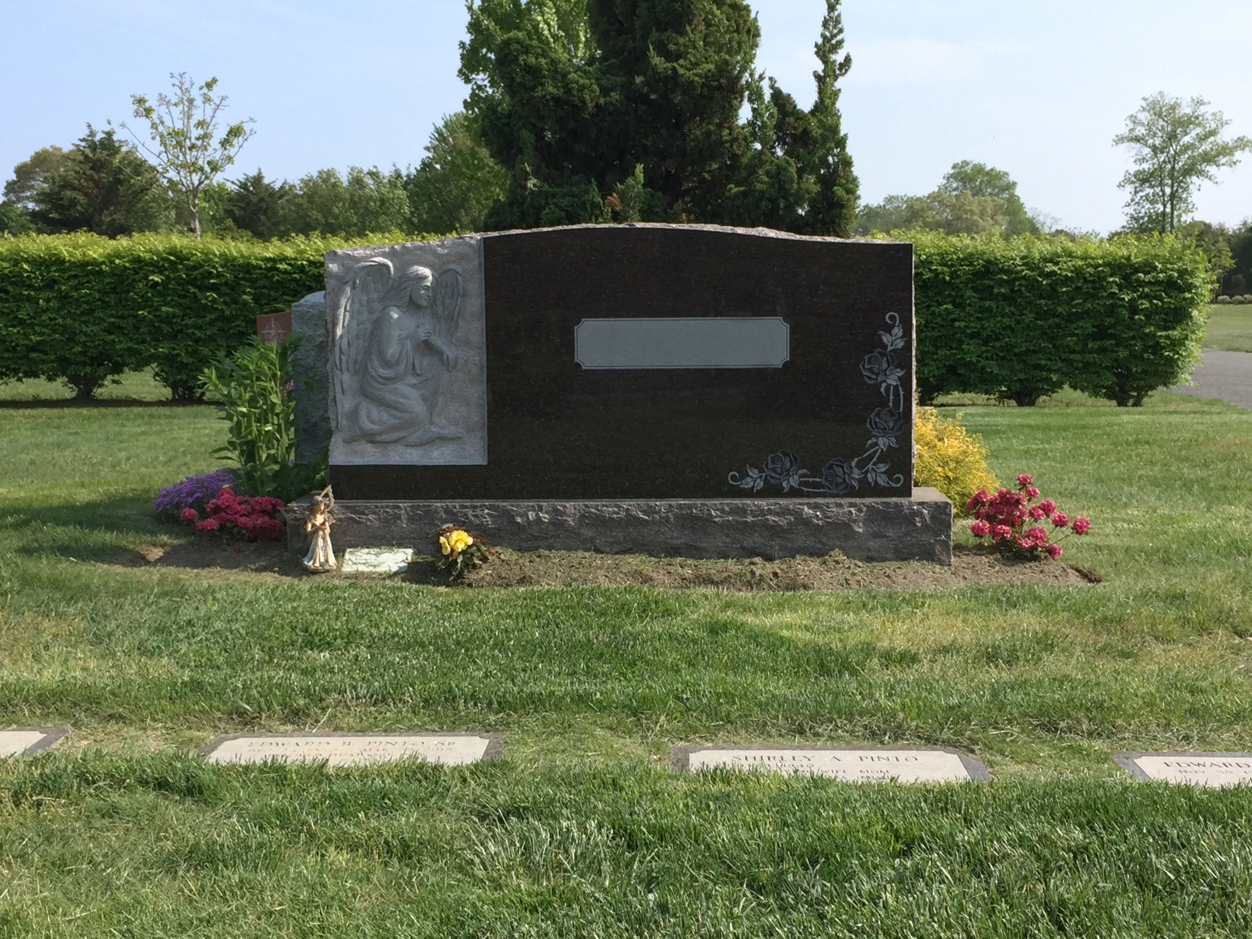 A cemetery scene with granite gravestones, one featuring a carved angel with wings. The gravestones are surrounded by green grass, colorful flowers, and bushes, with trees and a clear sky in the background.