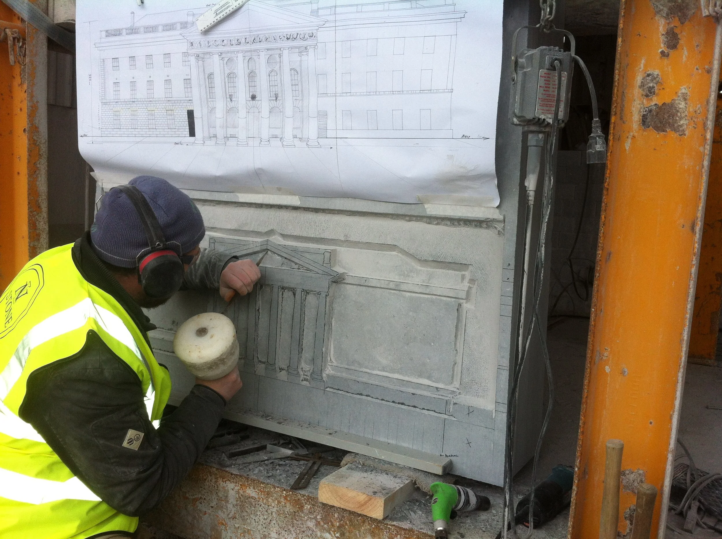 A worker in a yellow safety jacket and headphones is working on a stone sculpture, which appears to be an architectural detail like a column or frieze, with a blueprint of a classical building with columns and a pediment visible behind him.