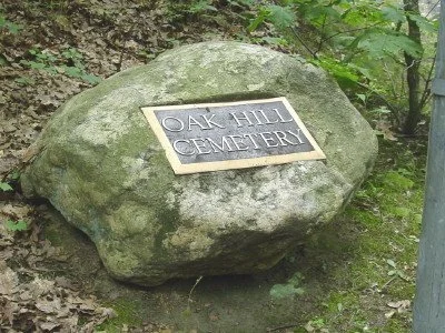 A large moss-covered rock with a metal plaque that reads 'Oak Hill Cemetery' in a wooded area.
