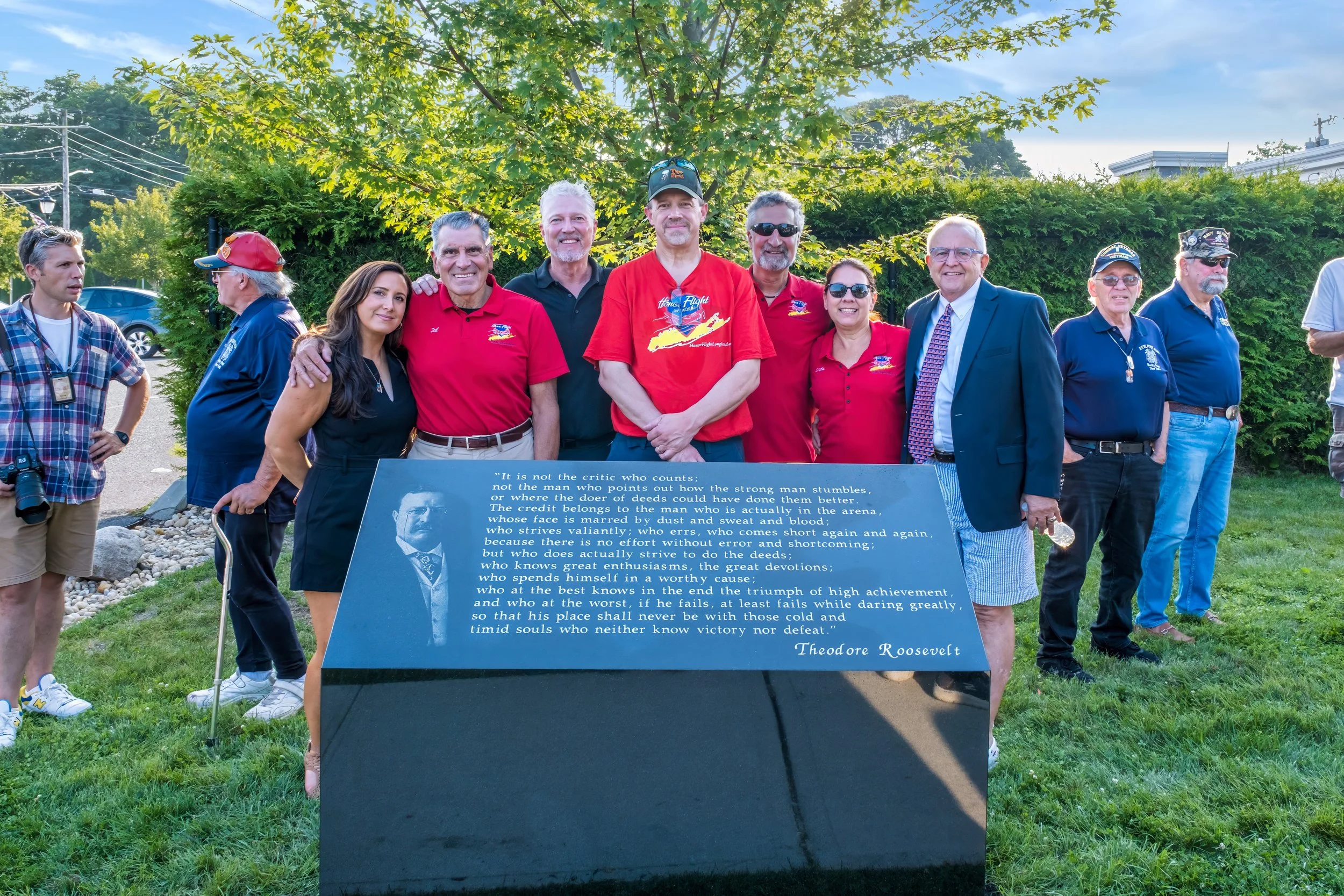Group of people standing outdoors in front of a large dark plaque with a quote and photo of Theodore Roosevelt, with a tree and blue sky in the background.