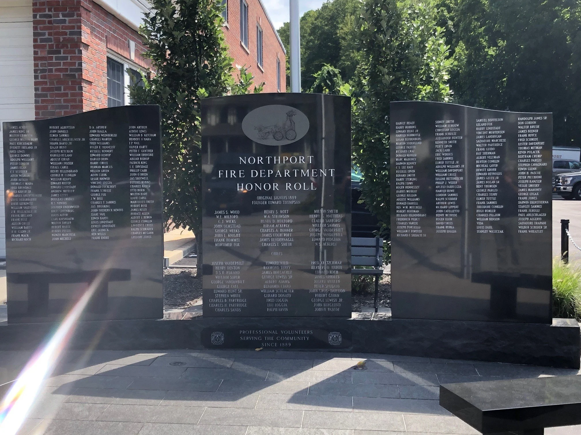 Memorial plaque dedicated to the Northport Fire Department Honor Roll, listing names of firefighters and volunteers since 1889, in front of a brick building with trees and a blue sky in the background.
