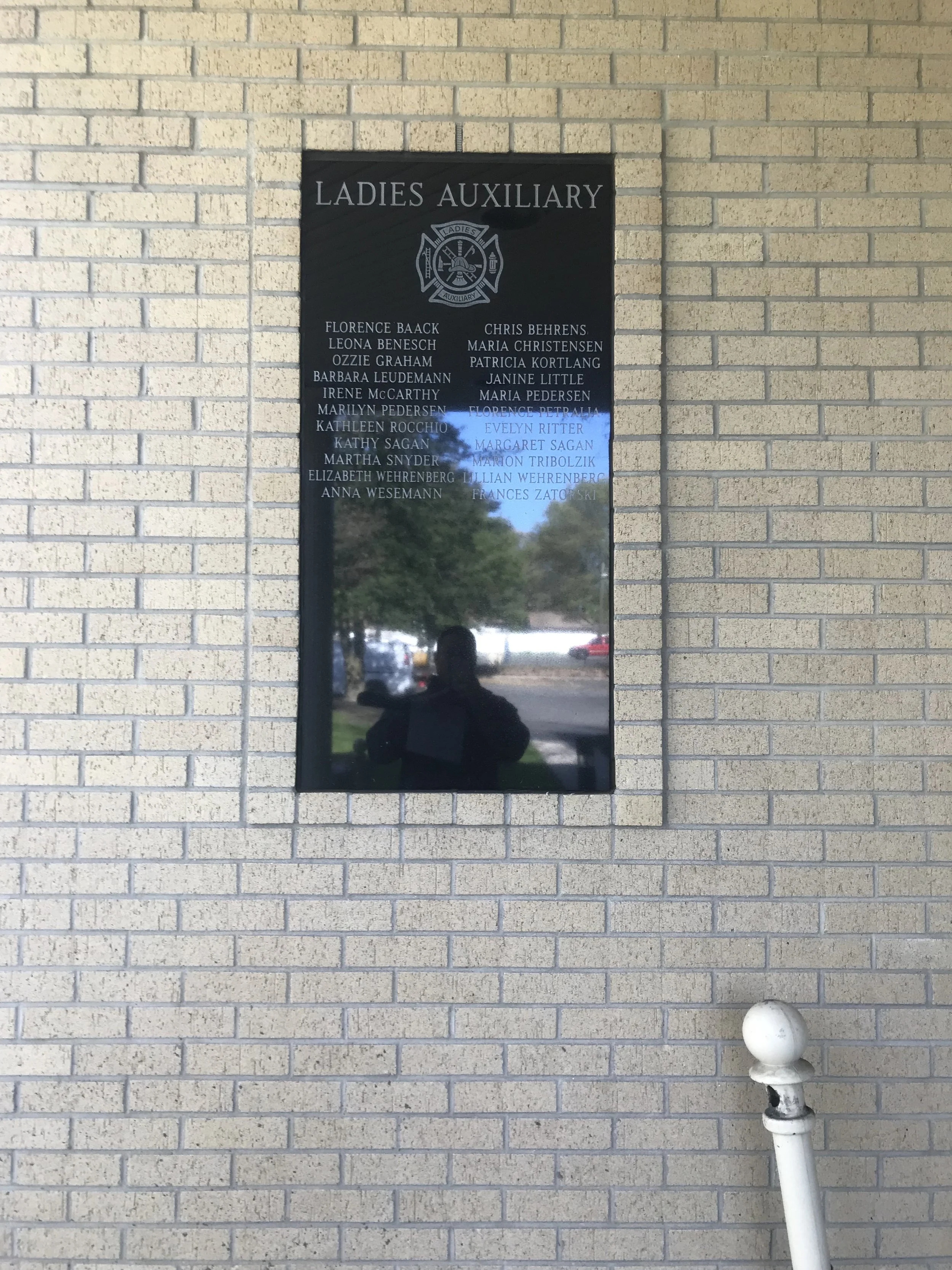 A reflective black sign on a beige brick wall with the title 'Ladies Auxiliary' and a list of names beneath. The reflection shows trees and a blue sky. A white bollard is in the foreground, leaning to the right.