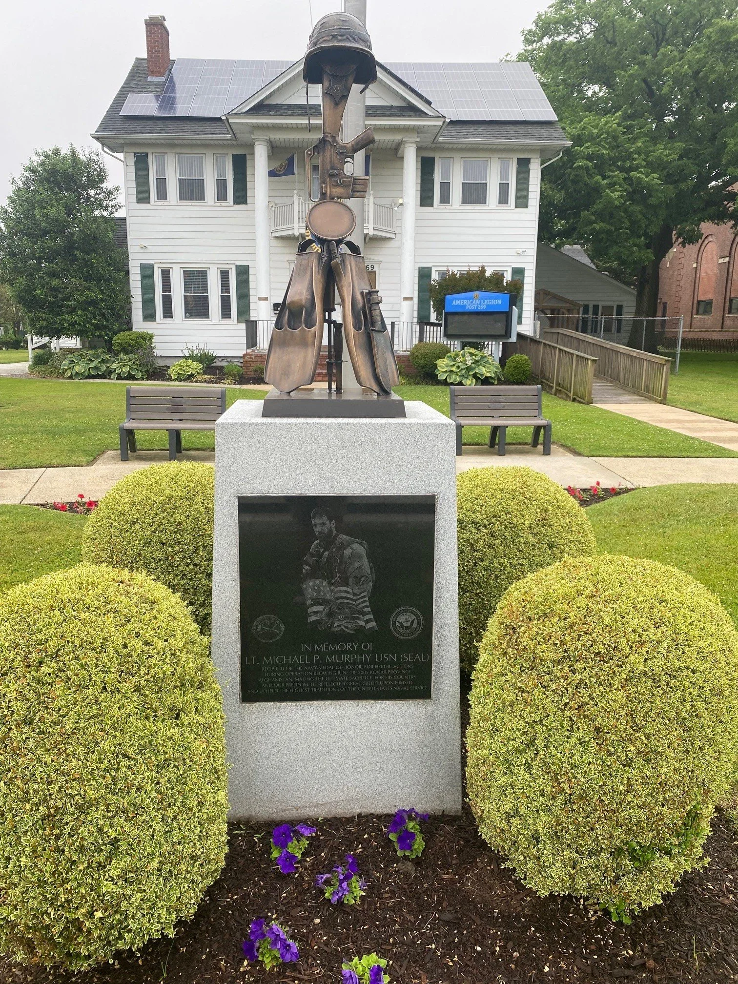 Memorial monument for Lt. Michael P. Murphy, USN, with a statue of a soldier's uniform and gear on a park lawn, surrounded by trimmed bushes and purple flowers, in front of a white house with solar panels.