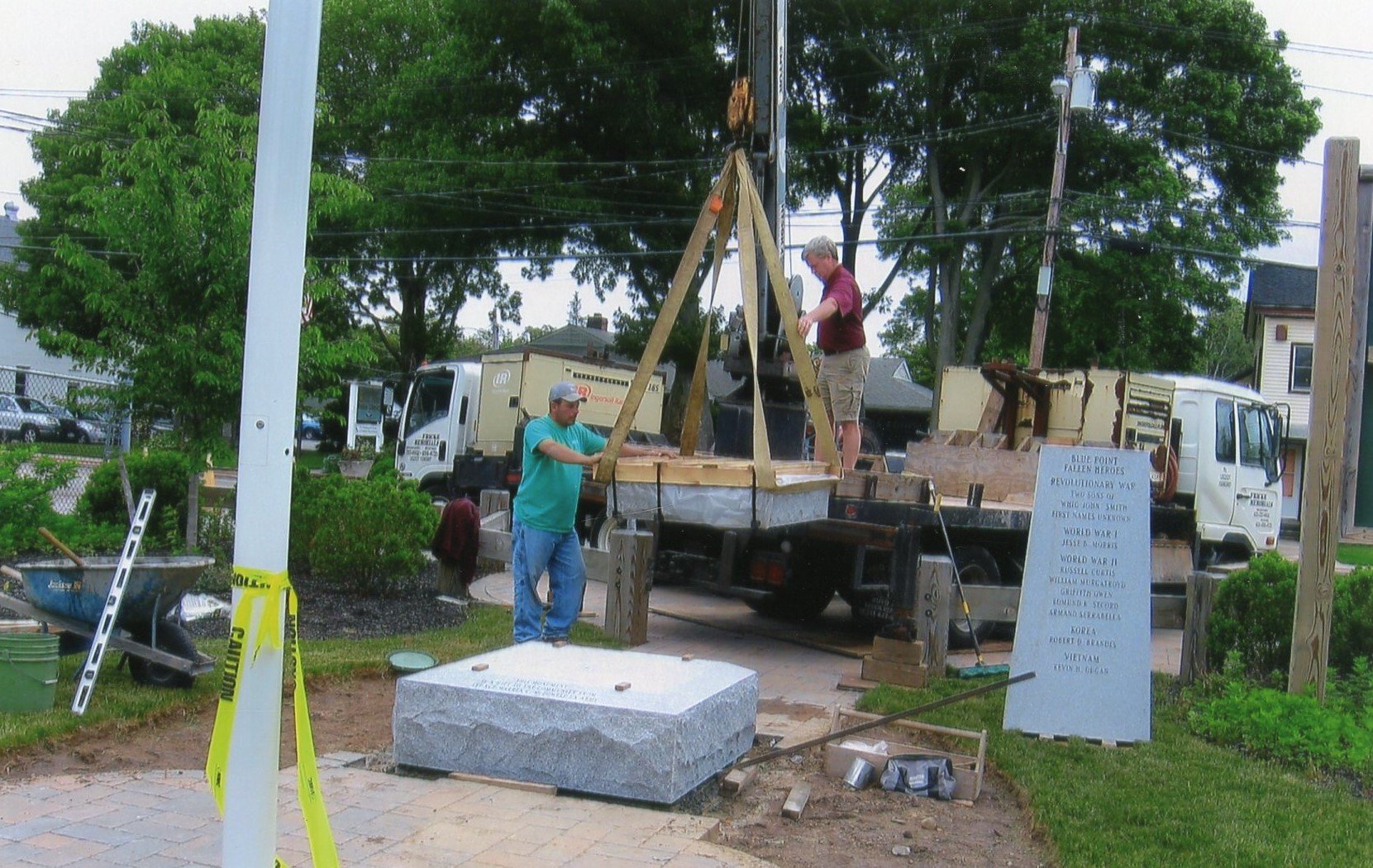 Two men are installing a large stone monument or plaque on a wooden platform using a crane. One man is standing on the platform, guiding the stone, while the other is standing beside it, helping. The scene takes place outdoors with trees, a white tru