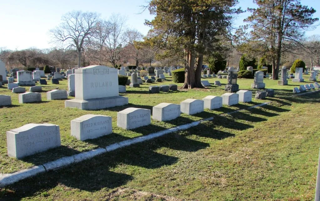 A cemetery with rows of gray tombstones, some shadows cast by trees, and leafless trees in the background.
