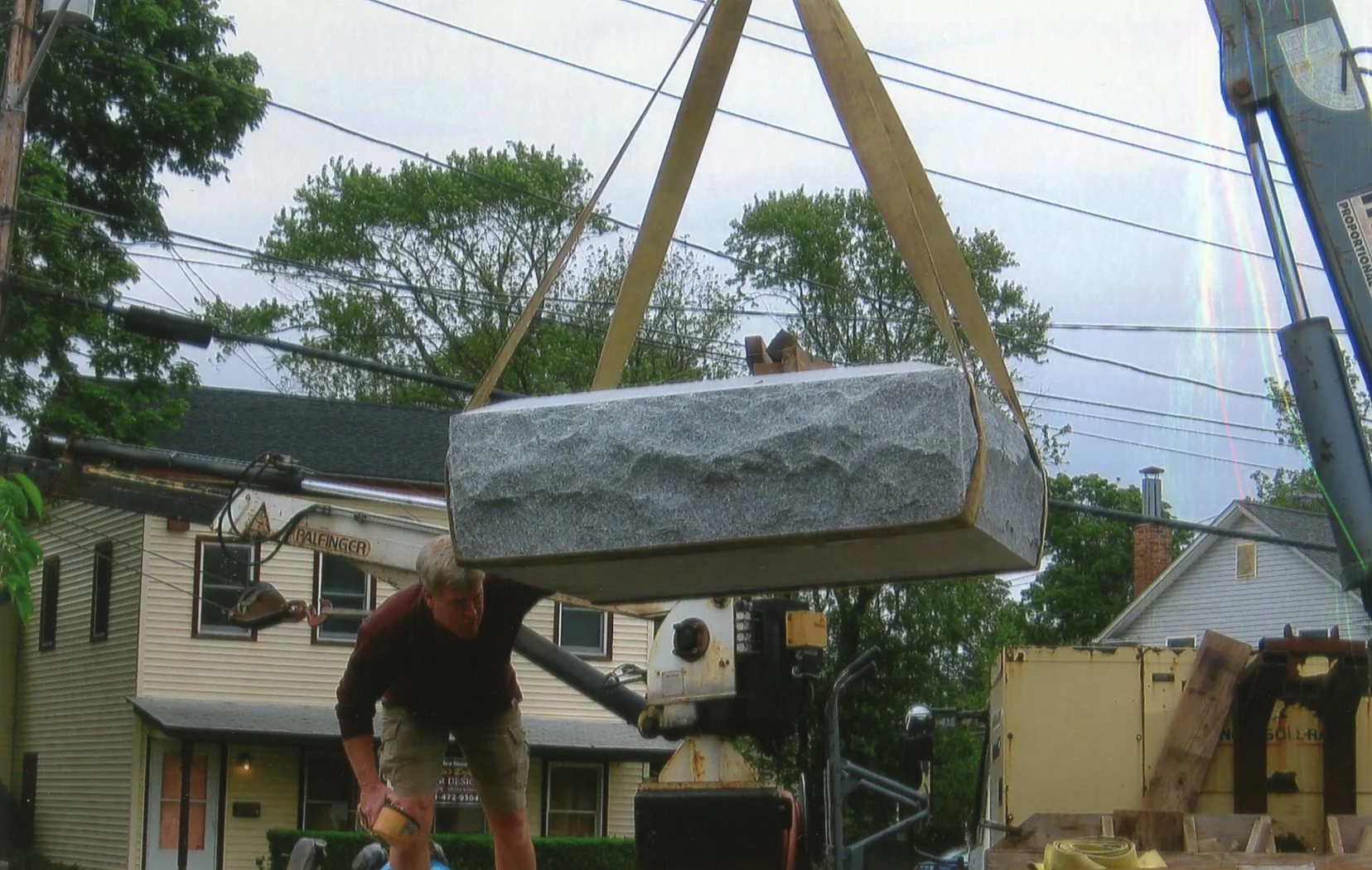 A large rock is being lifted by a crane into the air, with a man in a maroon shirt and shorts standing nearby, working on construction or transportation of the rock in a residential neighborhood with trees and houses in the background.