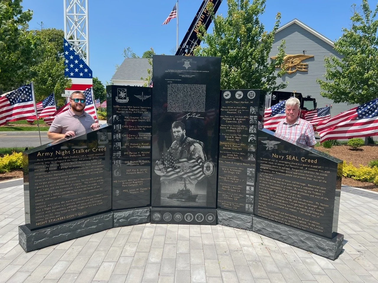 Two men standing on either side of a memorial wall dedicated to military service members, with American flags in the background, a building, and a clear blue sky.