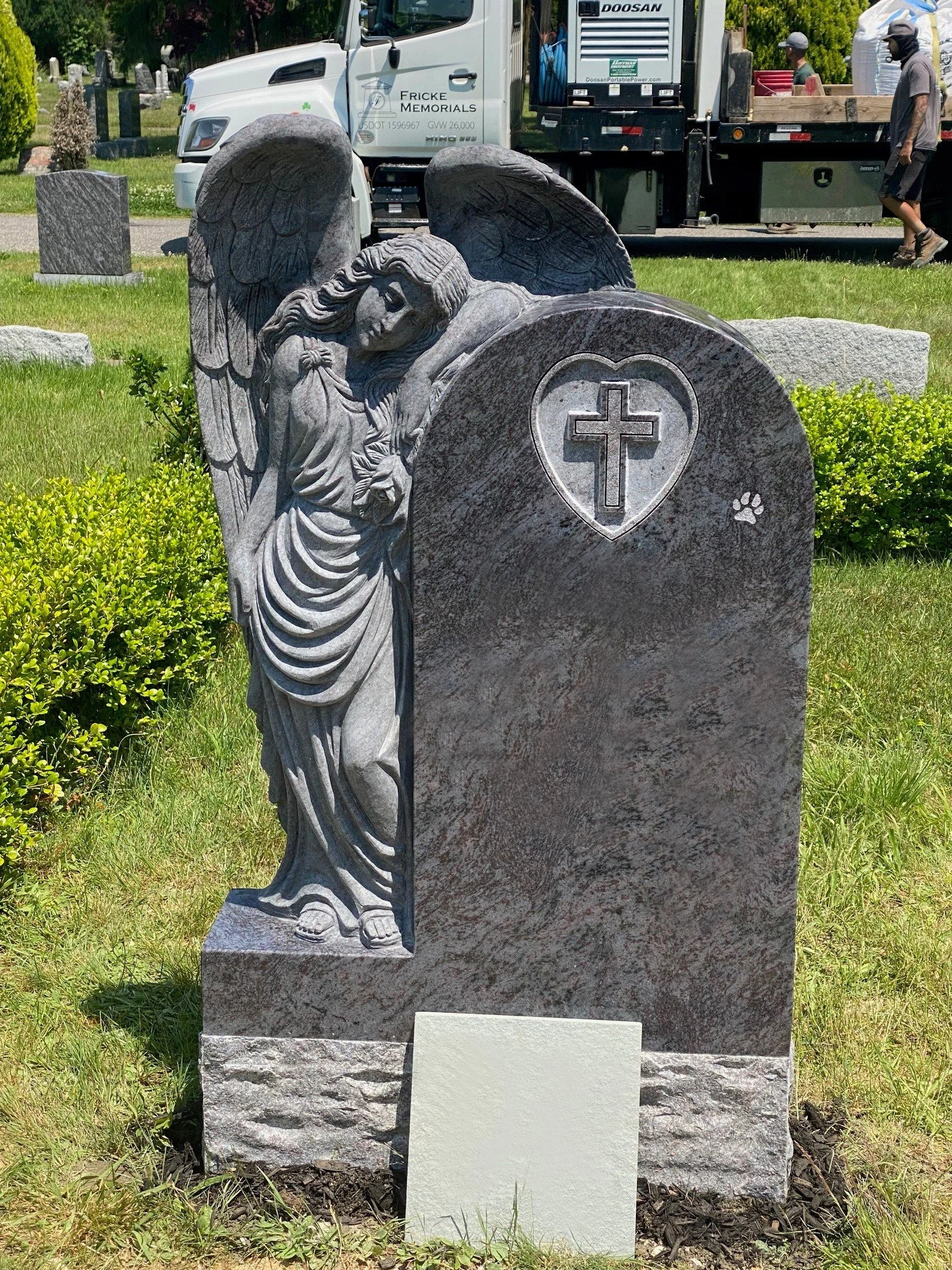 A cemetery grave marker featuring a detailed carved angel with wings and a sorrowful expression. The angel is holding a cloth and is depicted leaning on the gravestone, which has a heart with a cross carved into it. There is a paw print carved into the stone. In the background, there are trucks and people walking, and a grassy area surrounds the grave.