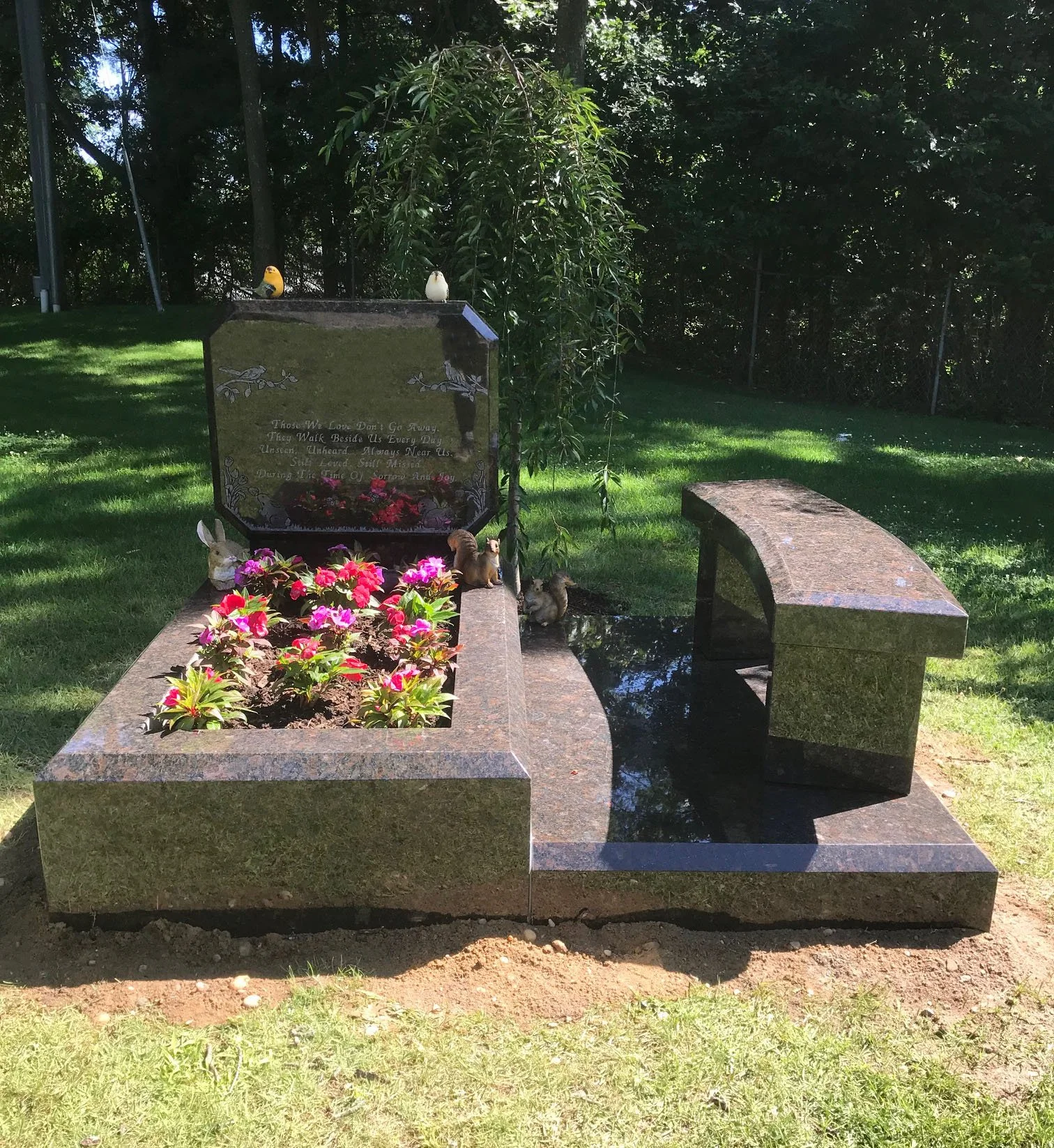 A grave site with a granite headstone and a curved bench in front. The headstone has an inscription, surrounded by pink flowers, and small animal figurines including a rabbit, squirrel, and birds. Two ceramic bird figurines are positioned on top of t