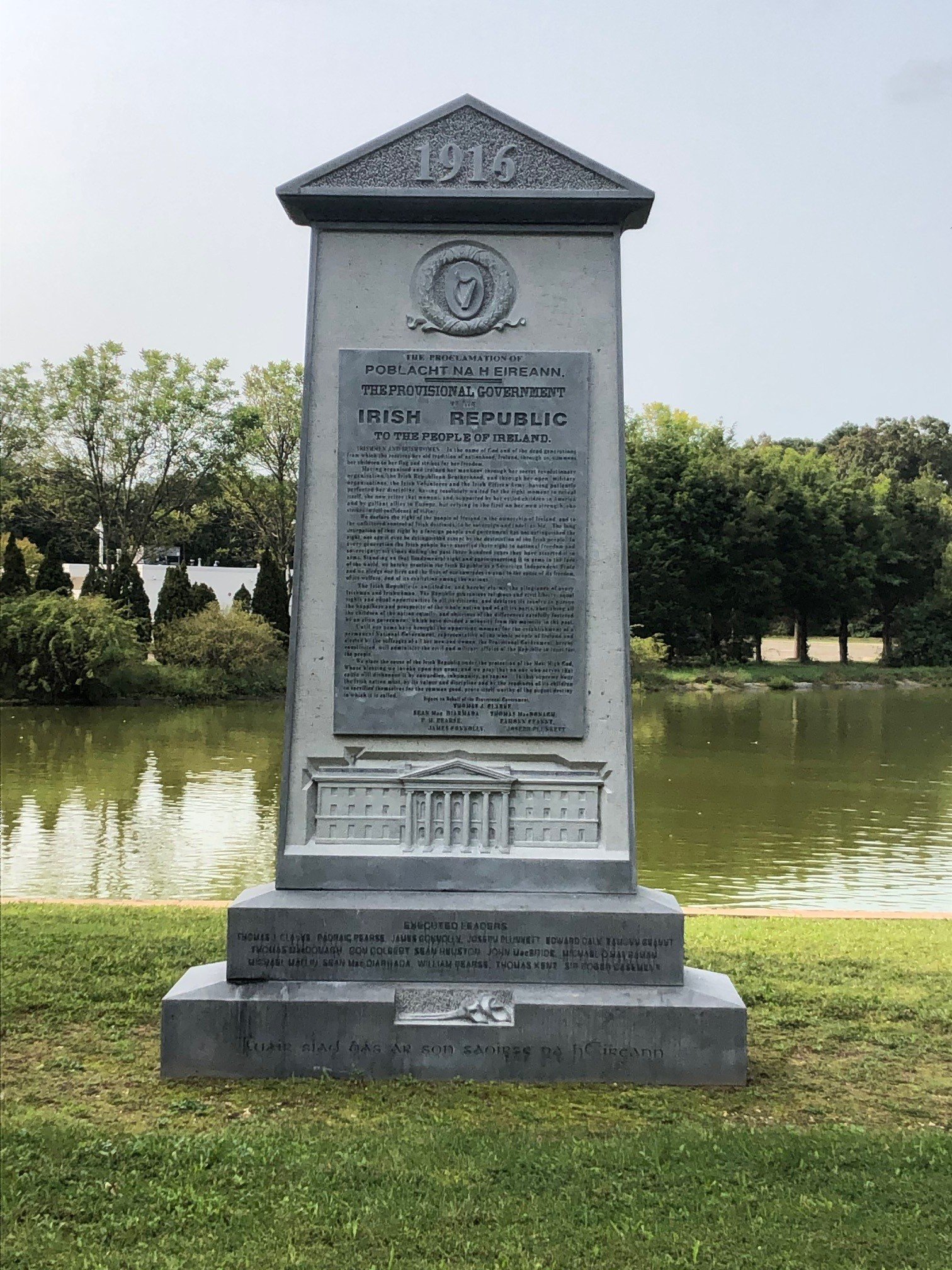 A stone monument with the year 1916 engraved at the top, located outdoors near a body of water with trees in the background. The monument contains inscriptions and a sculpted relief, commemorating Irish history or events.
