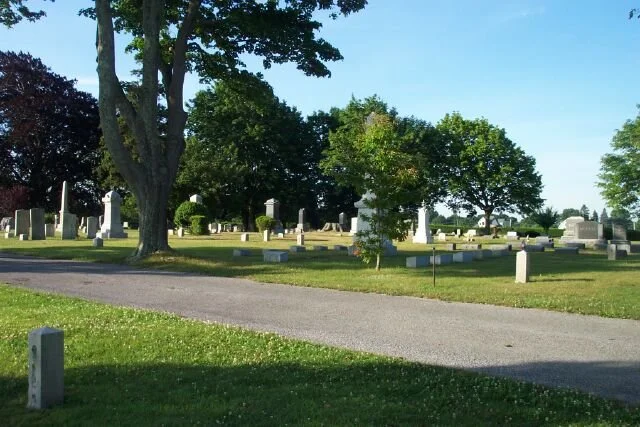 Wide view of cemetery with many different tombstones including older traditional obelisk monuments.  Green grass and trees with leaves in bloom.