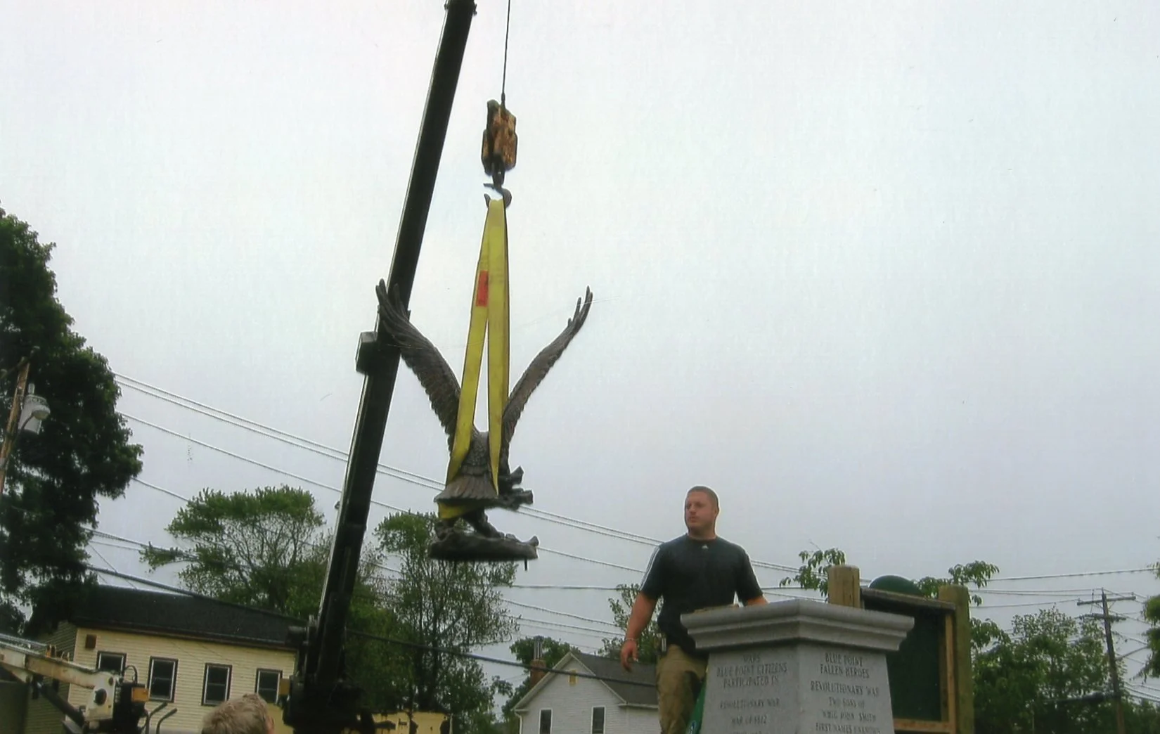 A man standing next to a monument or memorial, with a statue of an eagle being lifted by a crane during overcast weather.