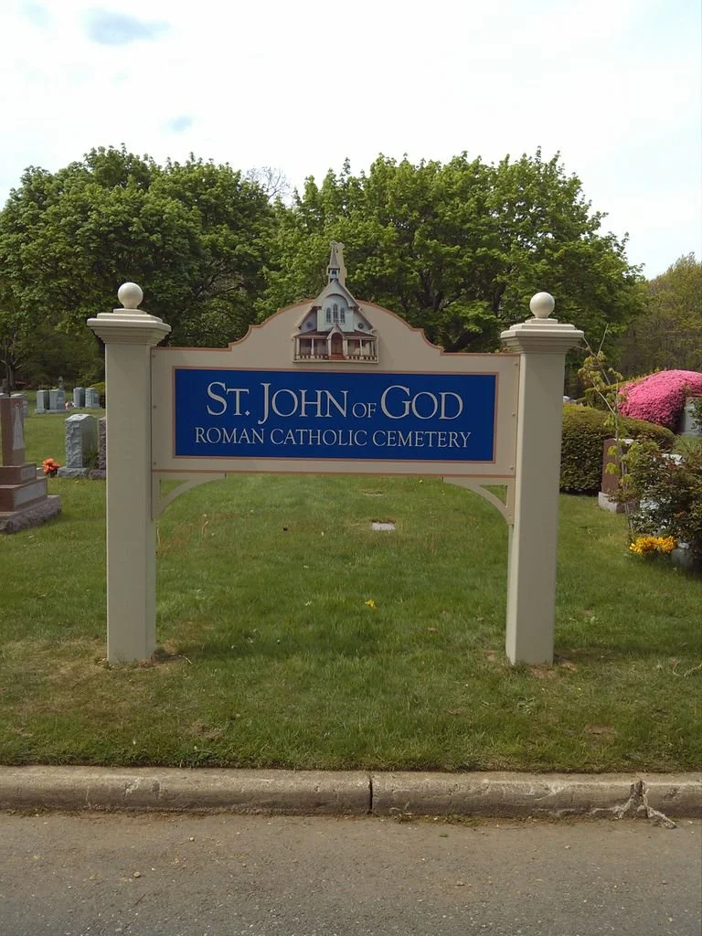 Sign for St. John of God Roman Catholic Cemetery with a small church illustration on top, in a grassy area with trees and flowers surrounding it.