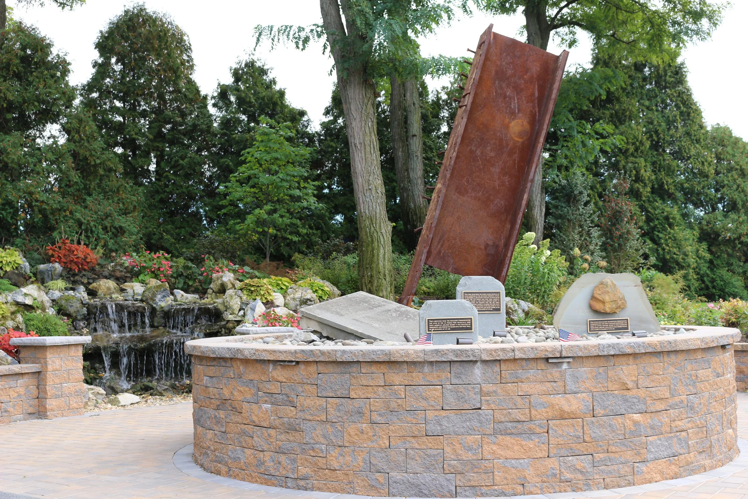 A memorial site with a curved brick wall featuring engraved plaques, a rusted metal structure about 8 feet tall, surrounded by trees, small waterfall, rocks, and flowering plants.