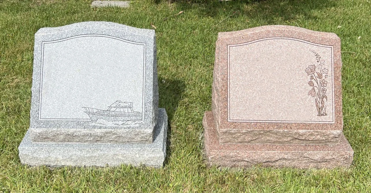 Two granite headstones on grass, one gray with a boat engraving, the other pink with floral engravings.