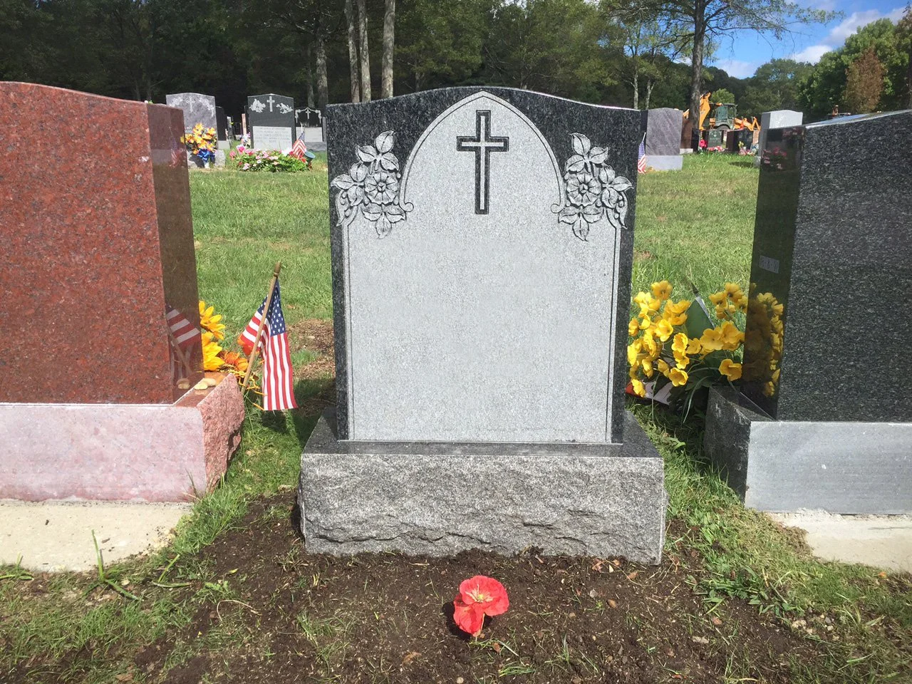 A gray granite tombstone with a cross and floral carvings at the top, in a cemetery with other graves and flowers around.