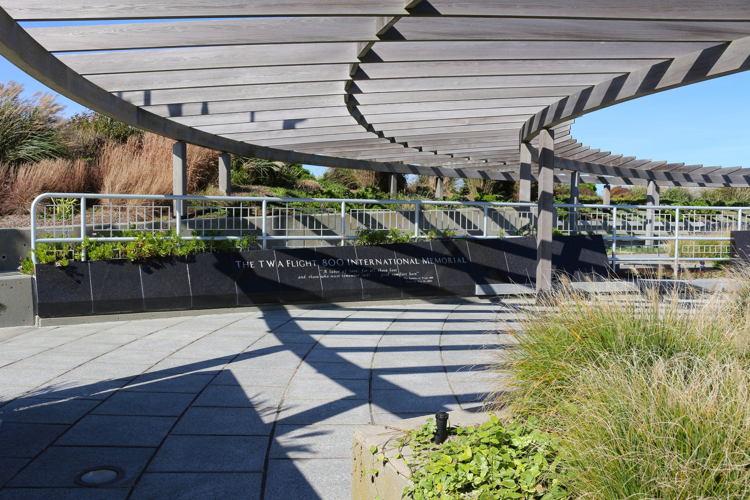 Memorial plaque at the Twin Flight 800 International Memorial, with a modern wooden canopy above, surrounded by landscaping and shadows.