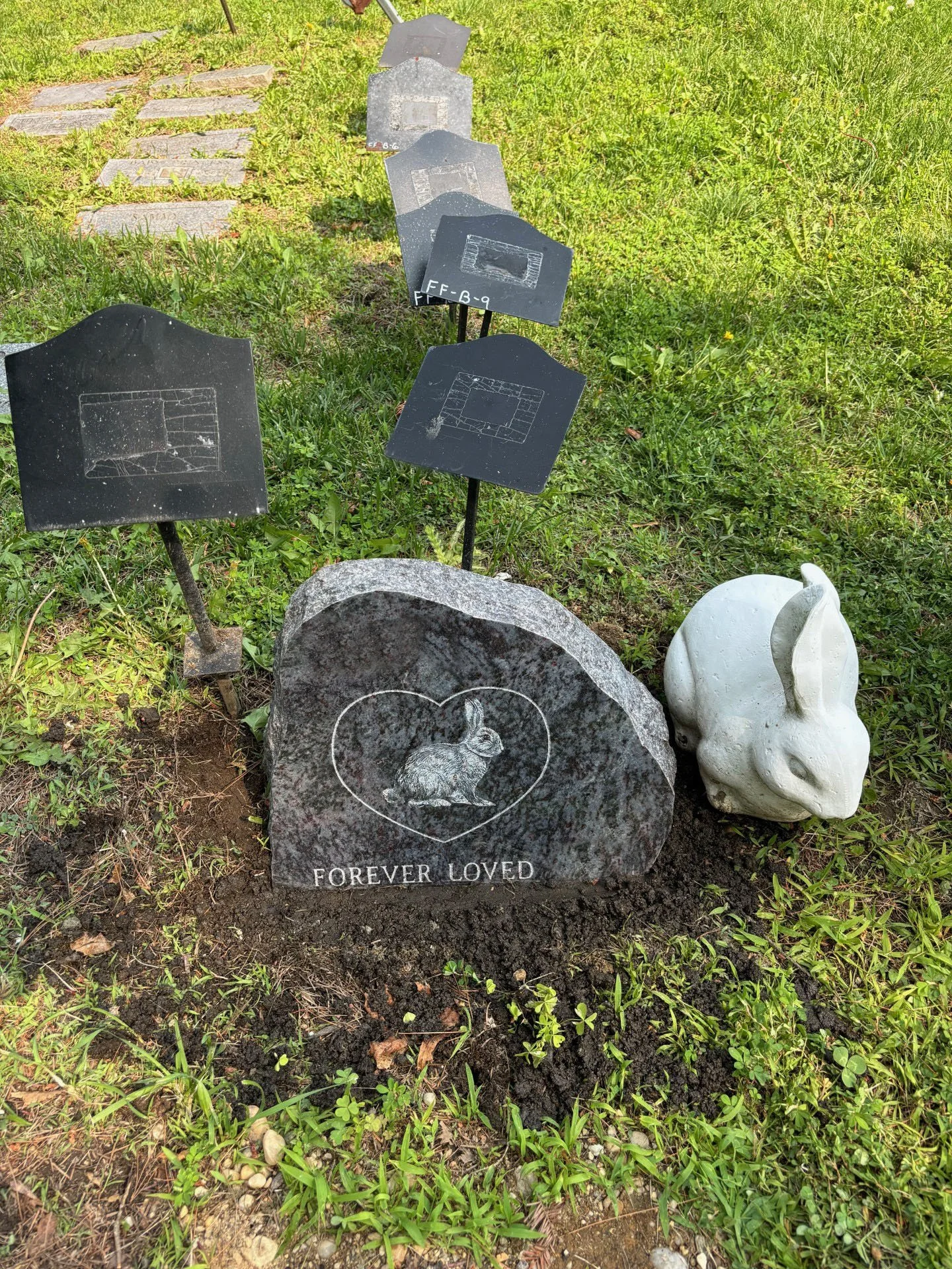 A small grave marker with a rabbit engraved on it and the words "Forever Loved" beneath. Surrounding the grave are multiple small black metal signs with sketches of gravestones, placed on the grass. There is a white rabbit-shaped decoration or statue