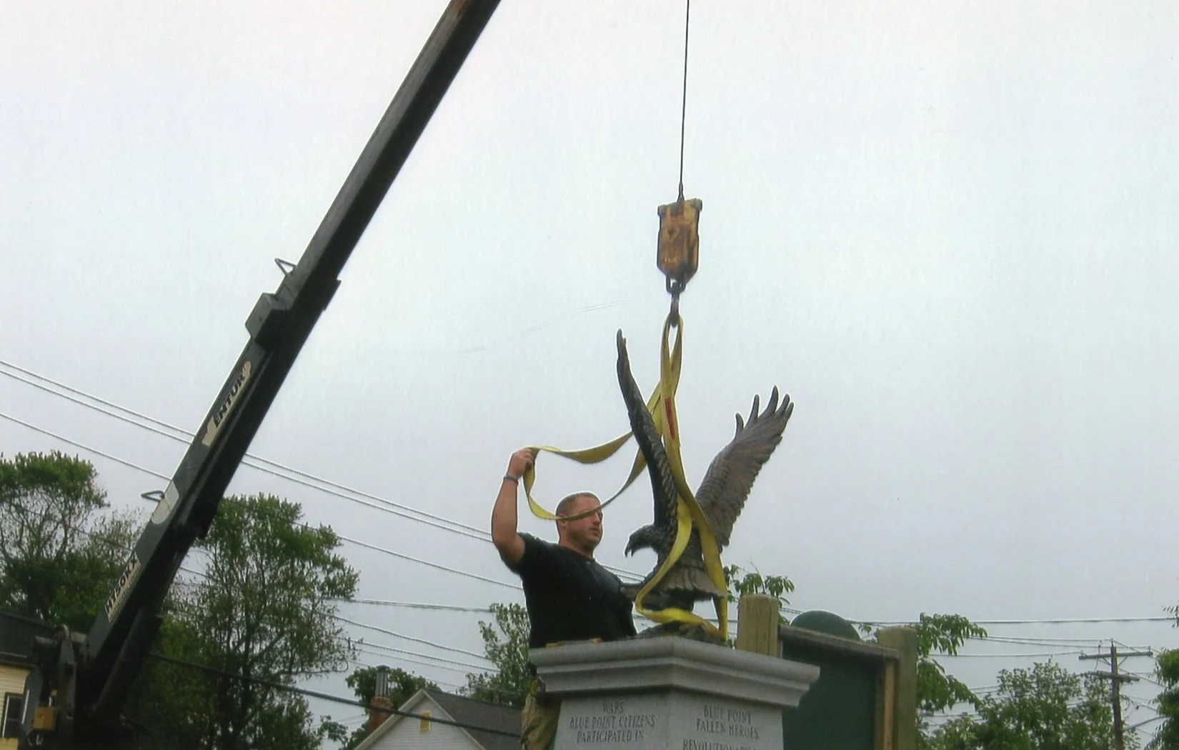 A man in a black shirt standing on a monument, holding a yellow strap attached to a crane, which is lifting a large eagle statue with outstretched wings. The sky is overcast, and there are trees and power lines in the background.