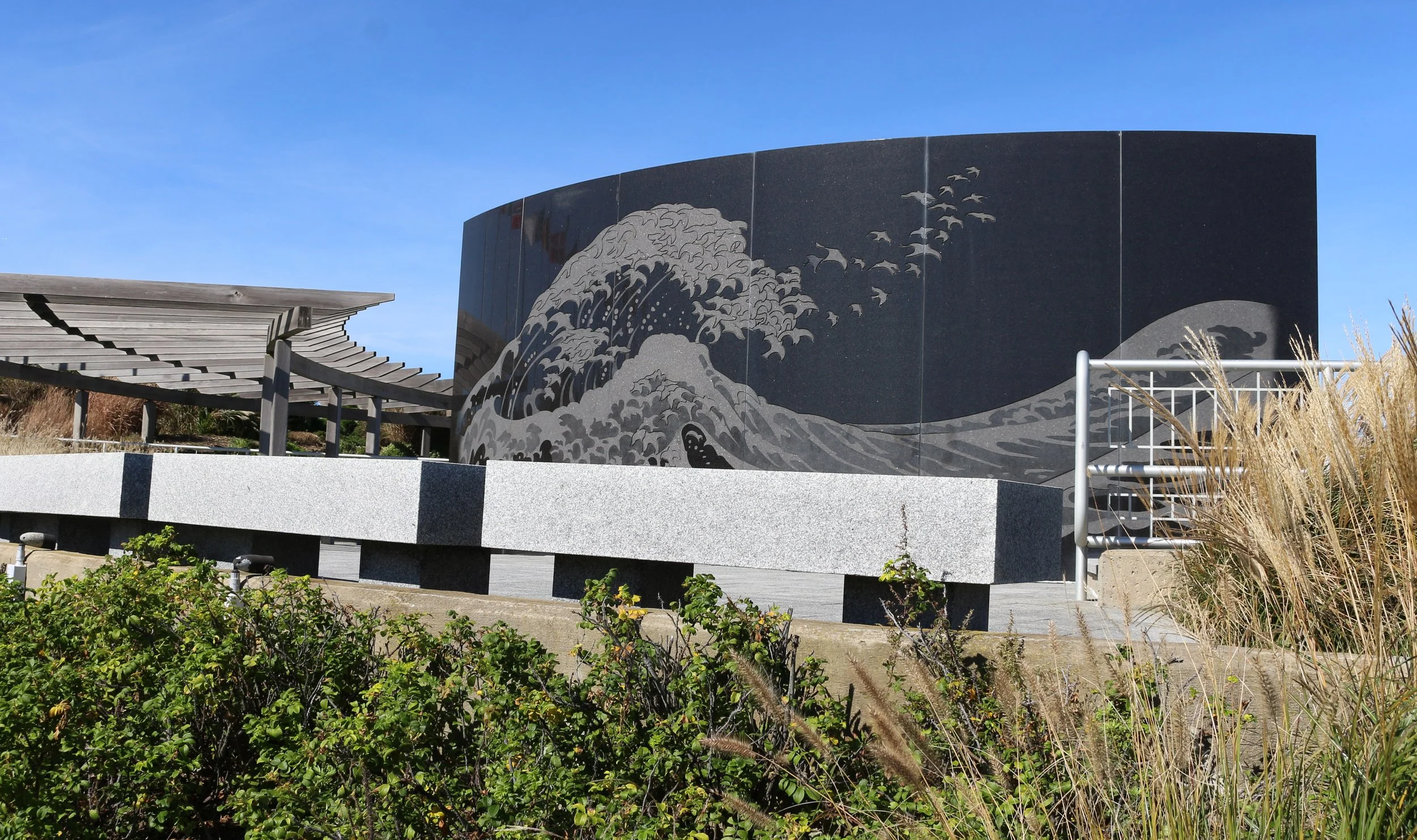 Black granite wall with a gray wave design and flying birds, part of an outdoor monument or art display with surrounding plants and a metal railing.