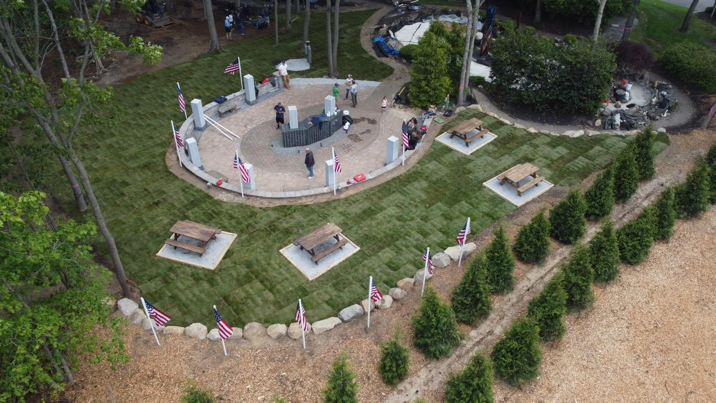 An aerial view of a small outdoor memorial area with American flags, picnic tables, and a circular monument surrounded by a walkway and trees.