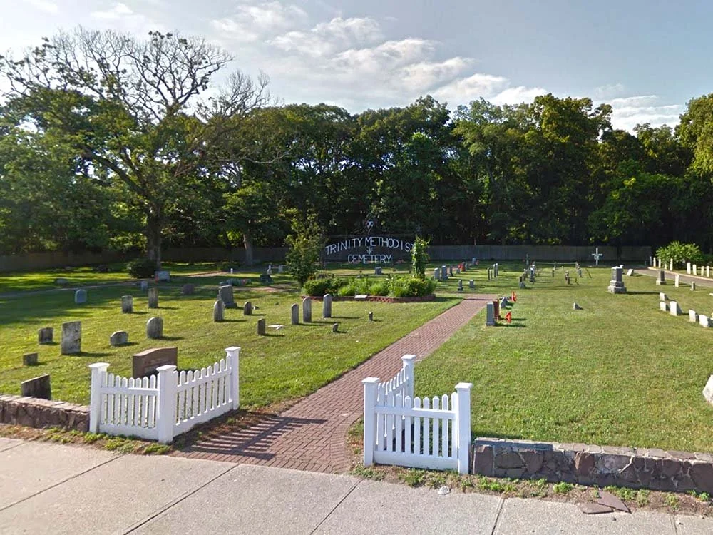Small white fence marks entrance to brick walkway into cemetery with old and new headstones lining each side.  Large cemetery sign at center.