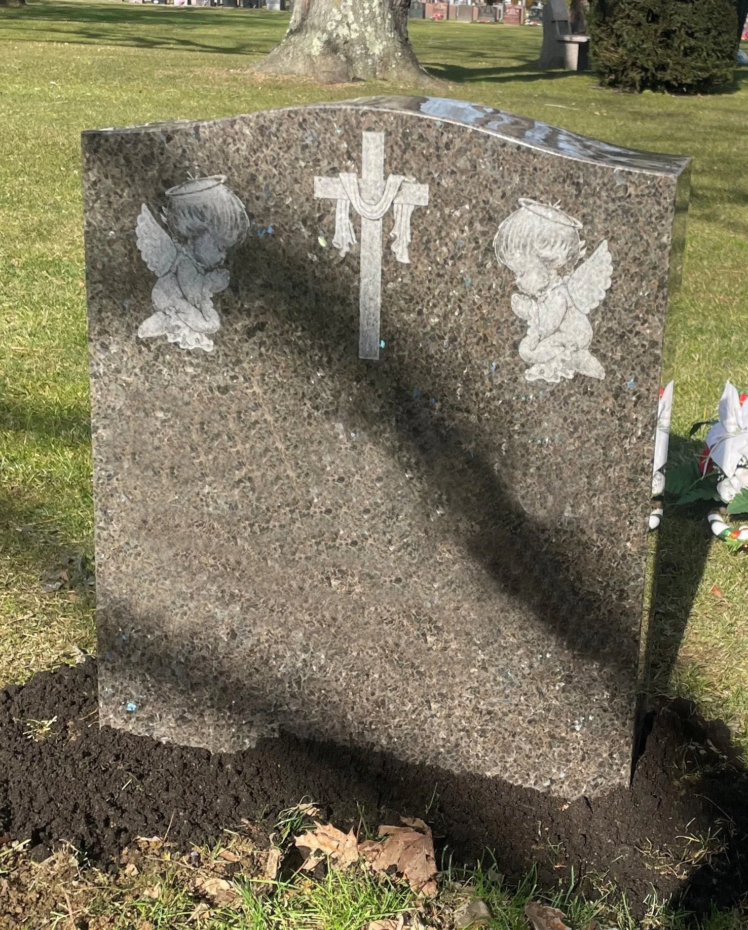 A polished granite headstone with engraved angels on either side and a cross with a draped cloth in the center, located in a grassy cemetery.