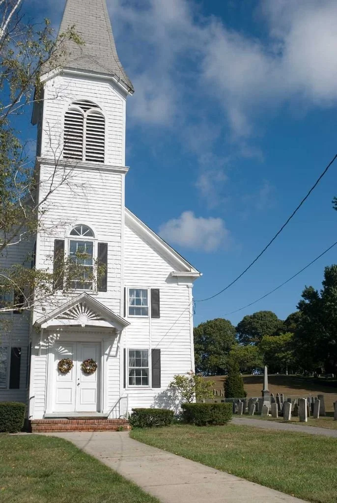 White church with cemetery headstones directly to the right of it.  Wreaths are on the double front door.