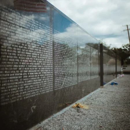 Photo of a black memorial wall with inscribed names and text, reflecting the cloudy sky and street in the background.