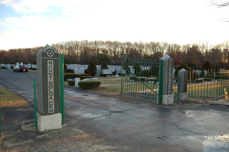 A cemetery entrance with a sign that reads 'Mount Golda Cemetery' and a gate, with rows of headstones and graves in the background.