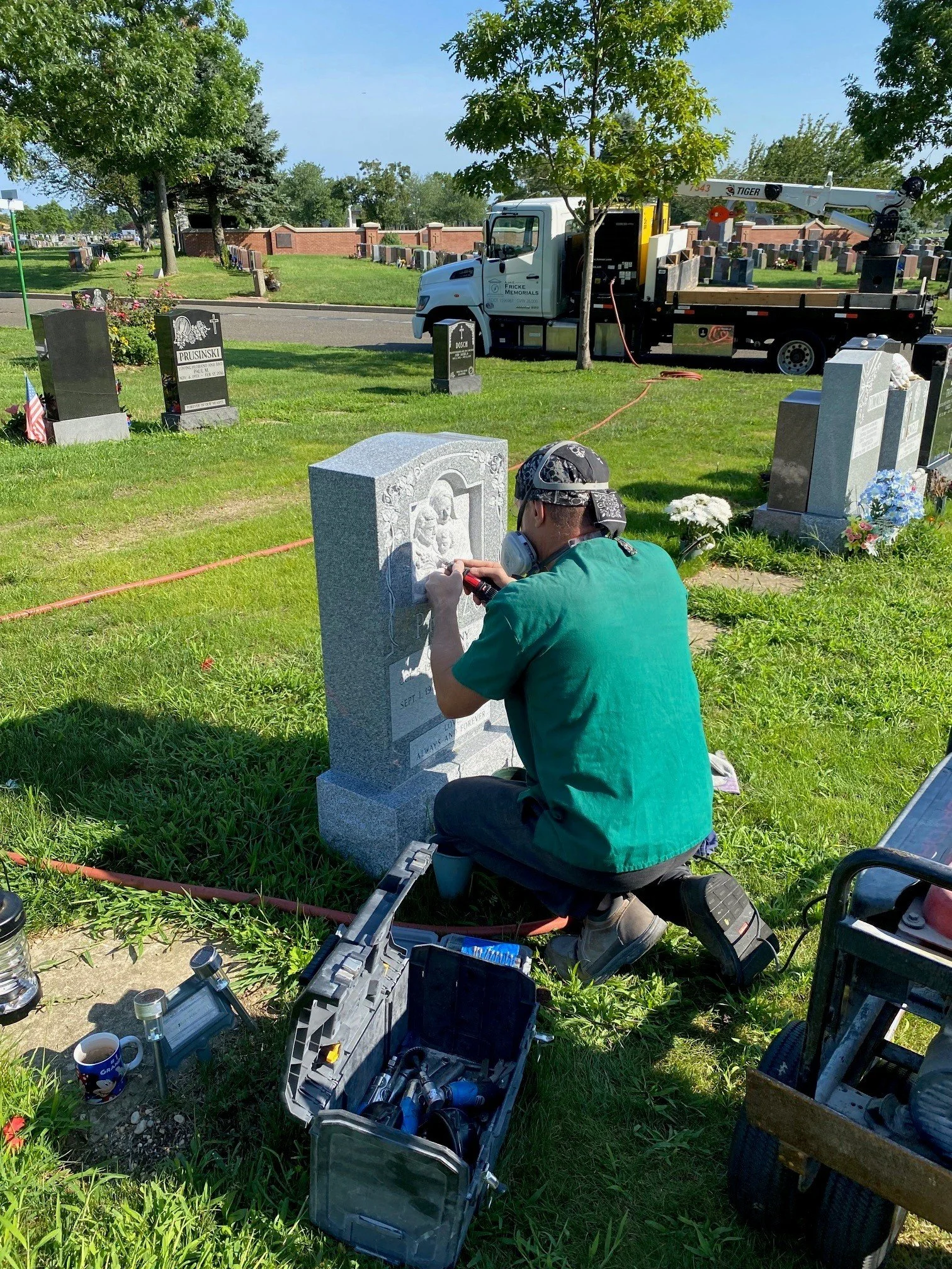 A man working on a gravestone in a cemetery, with tools and equipment around him. A truck with a crane is parked nearby.