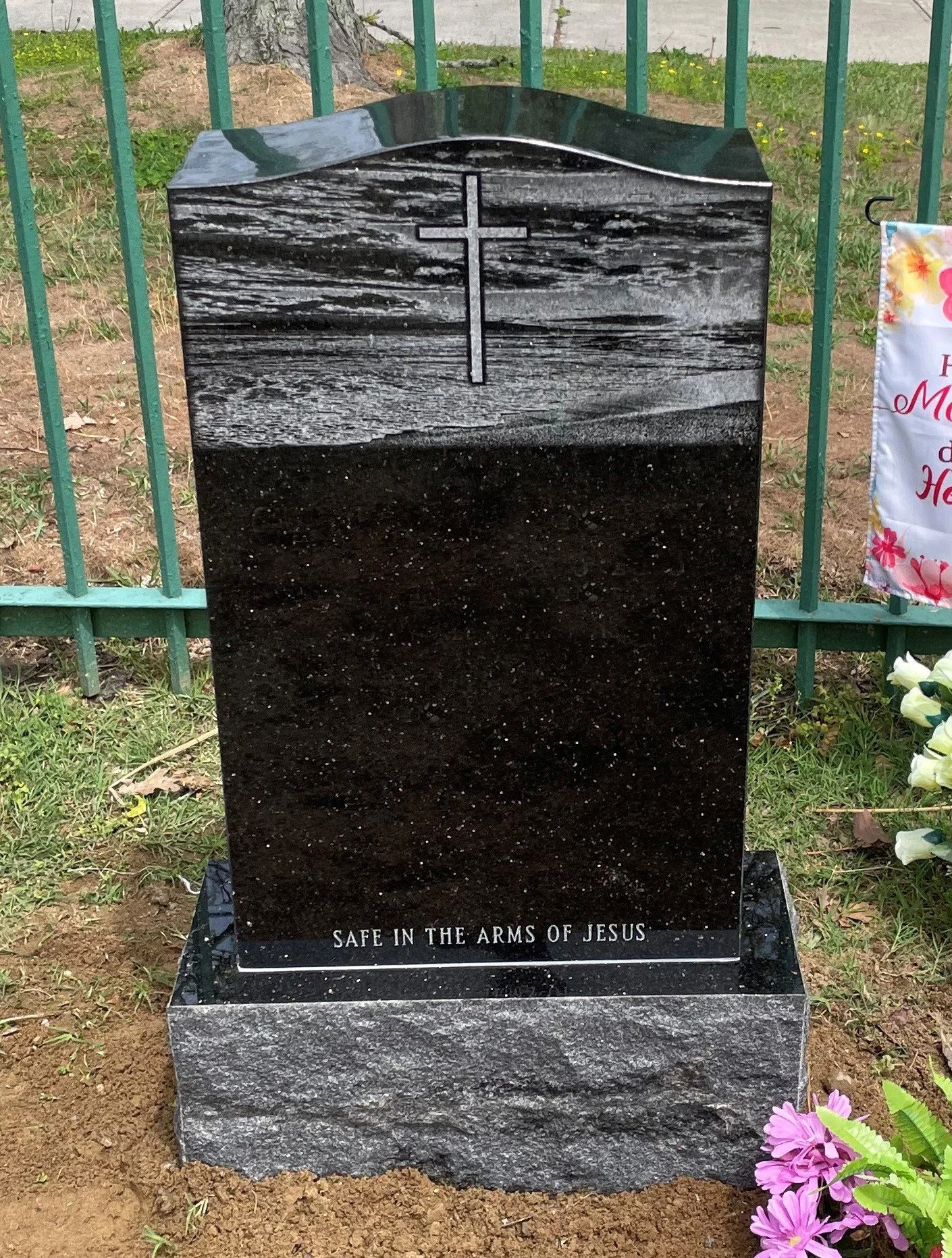 Black polished headstone with a carved cross at the top and engraved inscription at the bottom that reads "Safe in the arms of Jesus". The headstone is surrounded by flowers and greenery, indicating it is in a cemetery.