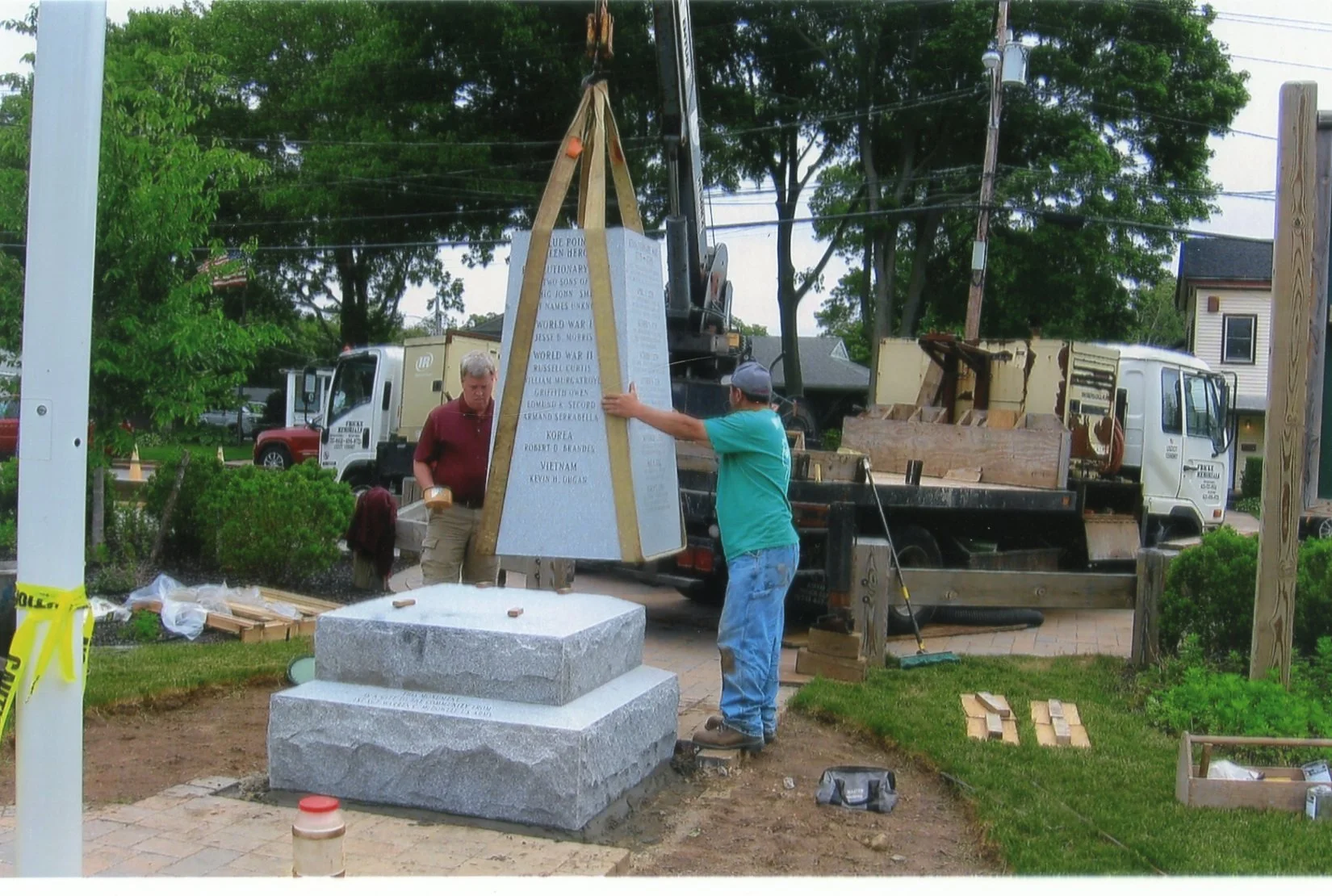 Two men are installing a memorial monument with an inscription in a yard, using a crane truck to lift the heavy stone piece.