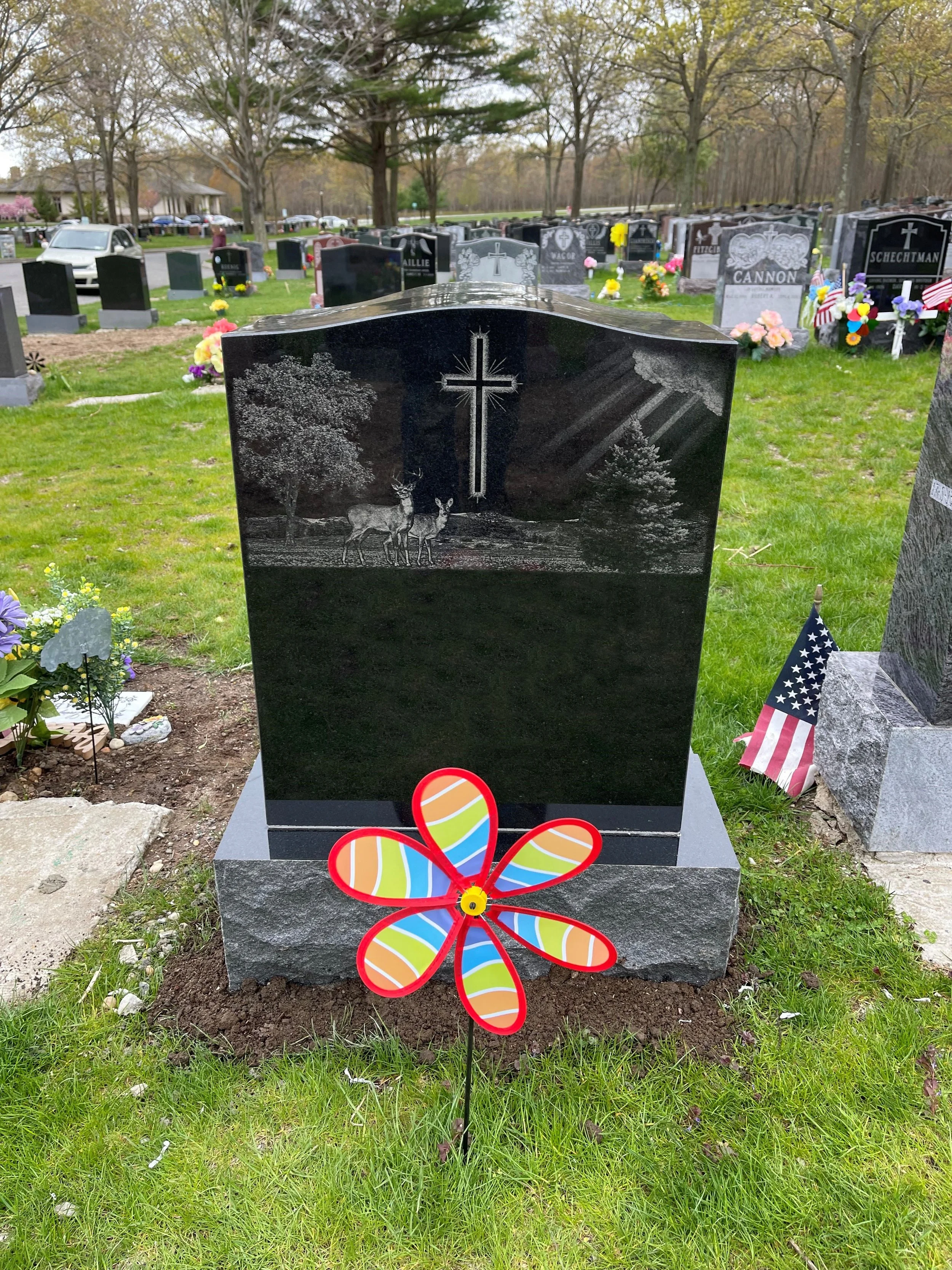 A black polished tombstone engraved with a cross, trees, and two deer etched into the surface. Flowers and American flags decorate the grave at a cemetery. A colorful plastic flower decoration is in front of the tombstone.
