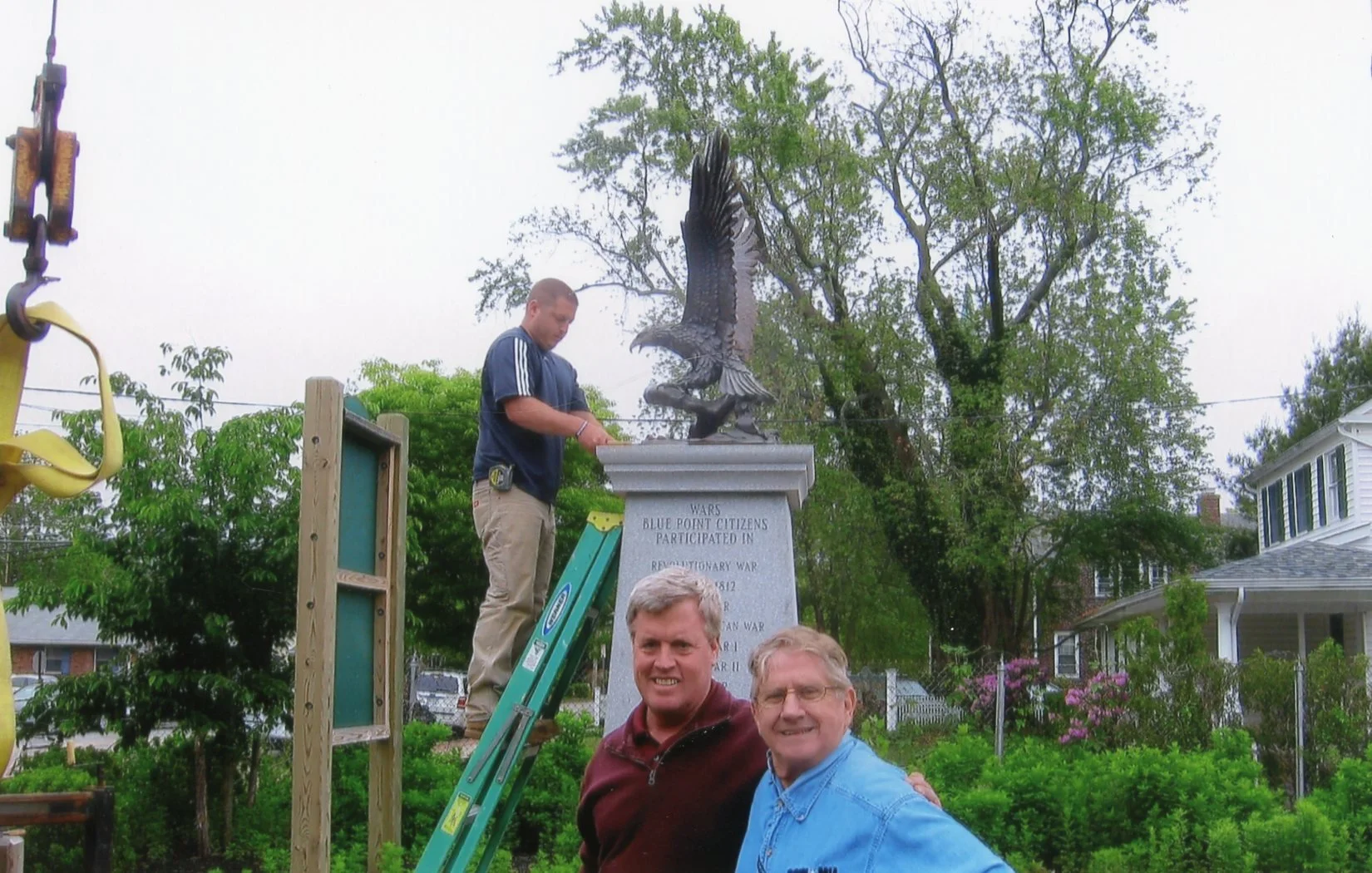 Two men standing in front of a memorial plaque with a large eagle sculpture on top, as a young man on a ladder adjusts or installs it. The memorial is surrounded by trees and houses, and the men are smiling at the camera.
