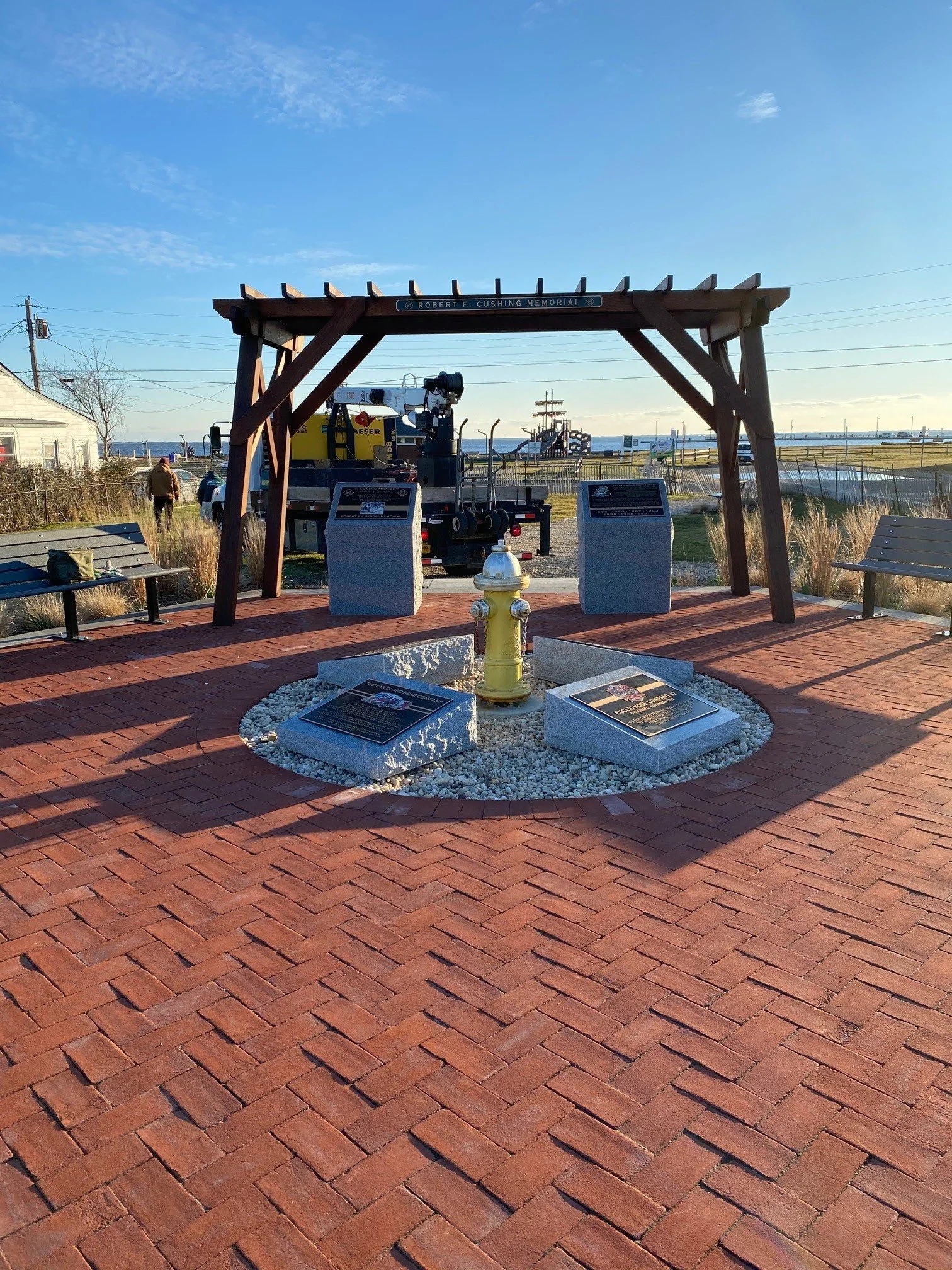 View of a coastal park memorial site with a wooden archway labeled 'Robert F. Cushing Memorial'. The site includes informational plaques, a yellow fire hydrant, benches, trees, and a view of the ocean in the background under a blue sky with some clou