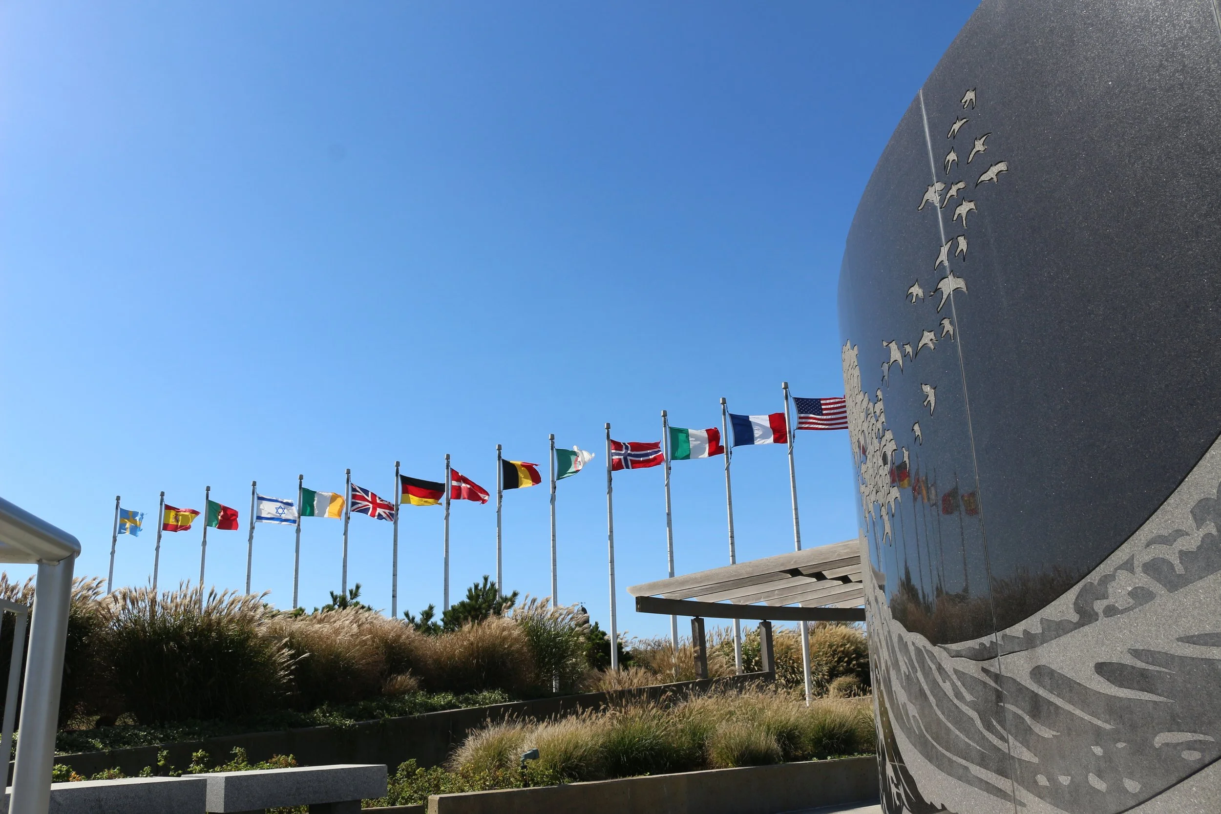 A row of international flags on flagpoles fly against a bright blue sky, beside a memorial or monument and some landscape with greenery and landscaping.