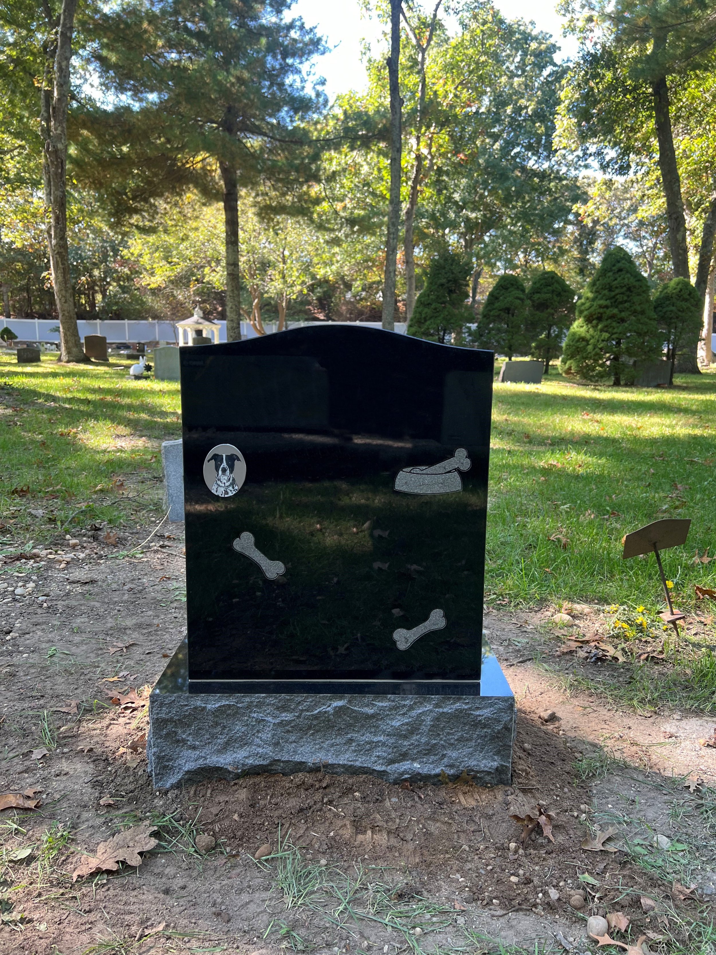 Black granite headstone with dog and bone engravings, located in a cemetery with trees and headstones in the background.