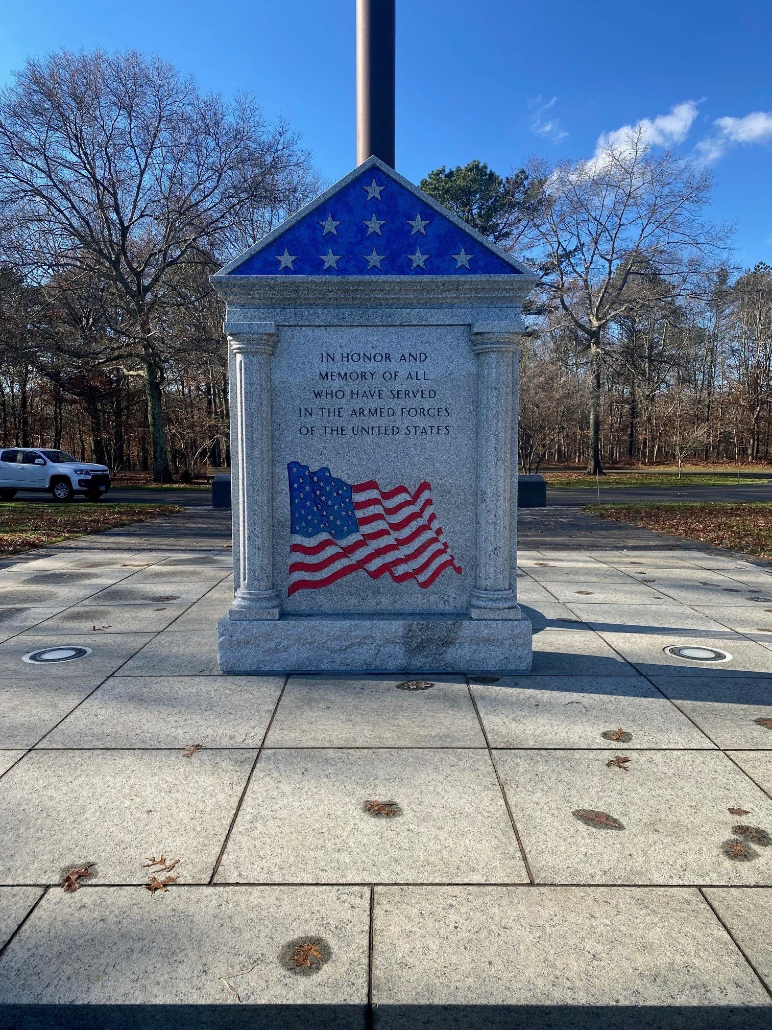 Memorial monument with a granite structure featuring an inscribed tribute, decorated with flags and star motifs, set outdoors on a paved area with trees and a car in the background.