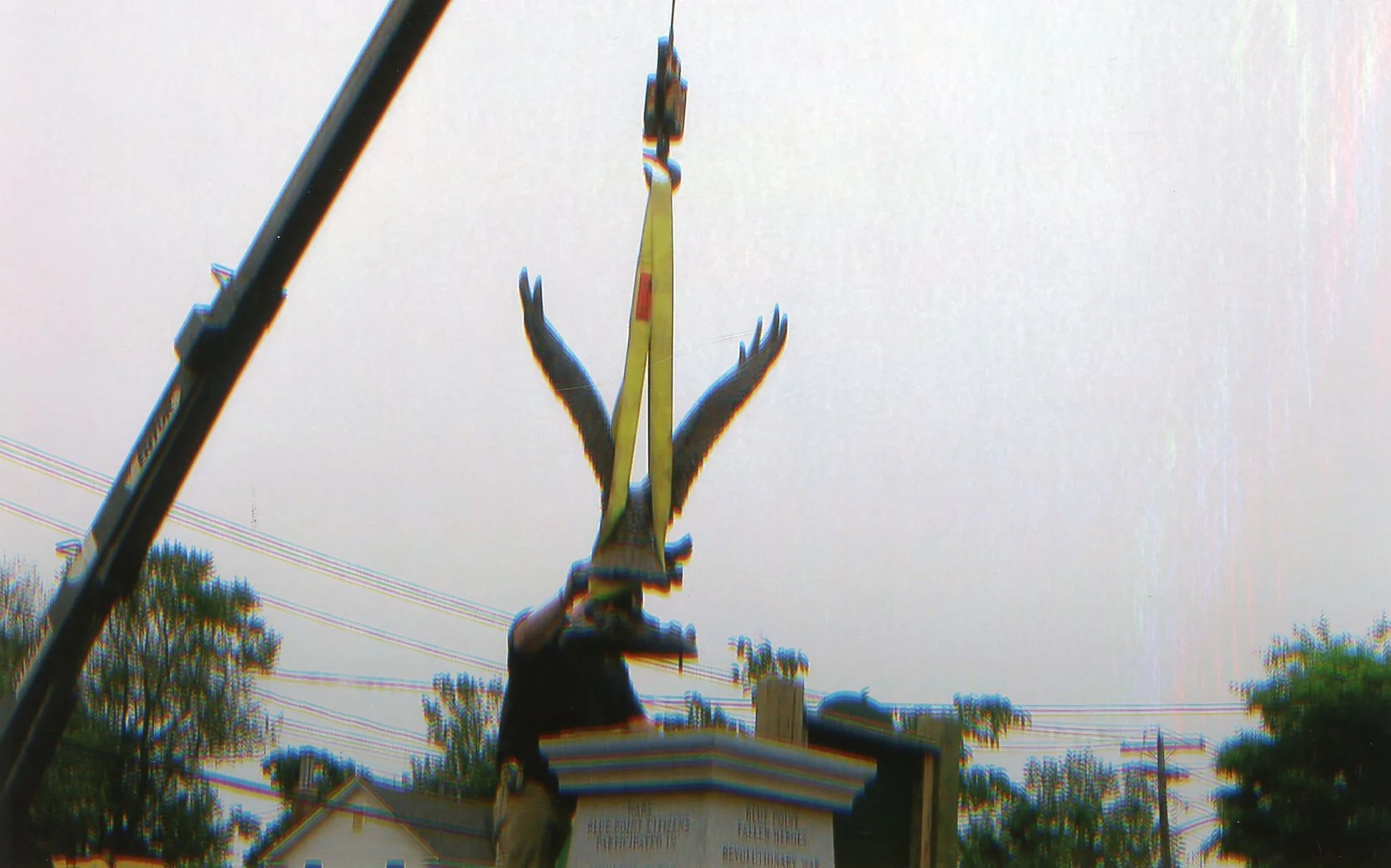 Person installing a large, gold-colored sculpture of a bird with outstretched wings on a wooden platform outdoors against a cloudy sky.