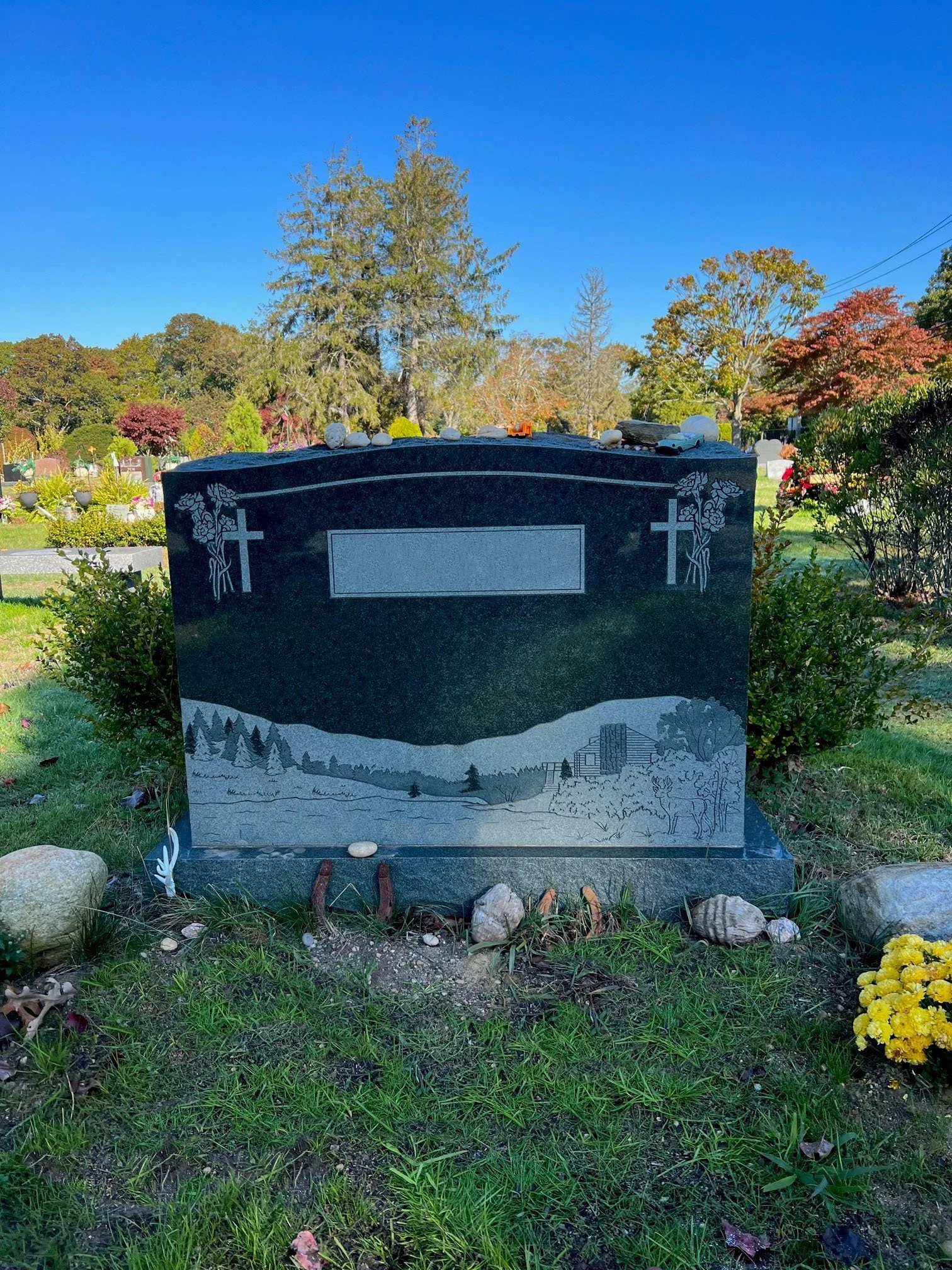 A polished black granite headstone in a cemetery with engraved roses at the top corners and a detailed landscape engraving at the bottom, surrounded by grass, rocks, and yellow flowers, with trees and a blue sky in the background.