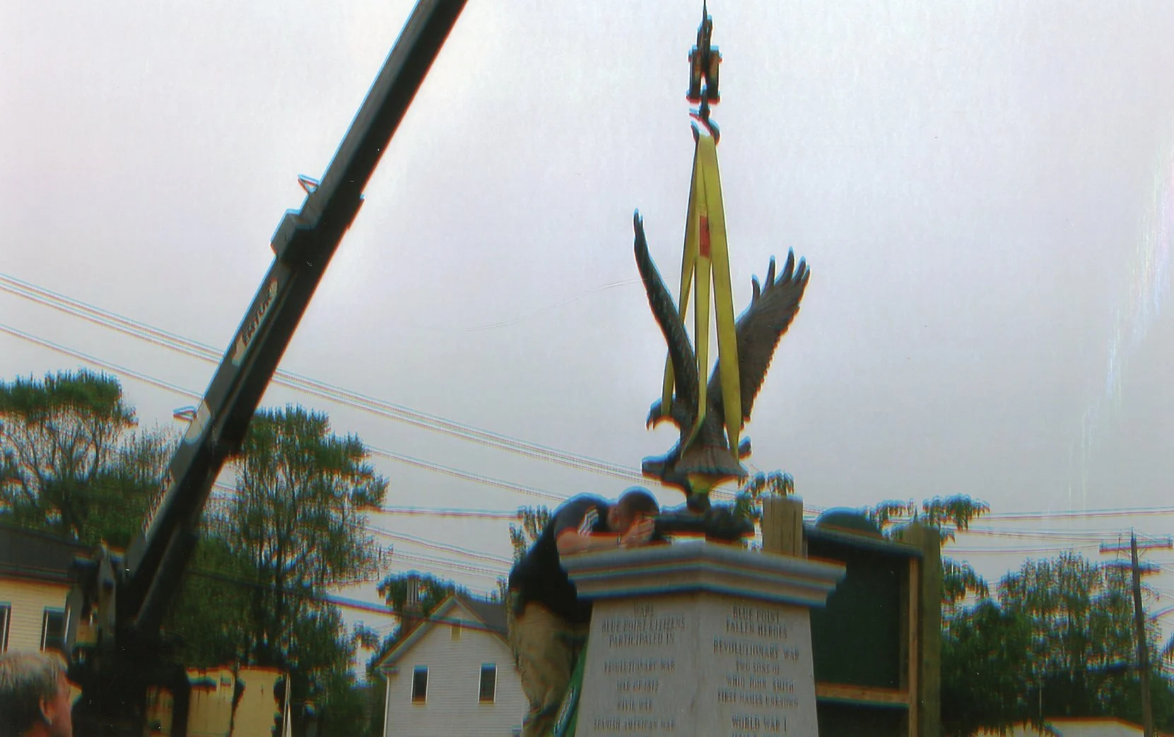 A man installing a large eagle statue on a monument with a crane. The monument has a plaque with inscriptions. Trees and houses are visible in the background, and utility lines run across the sky.